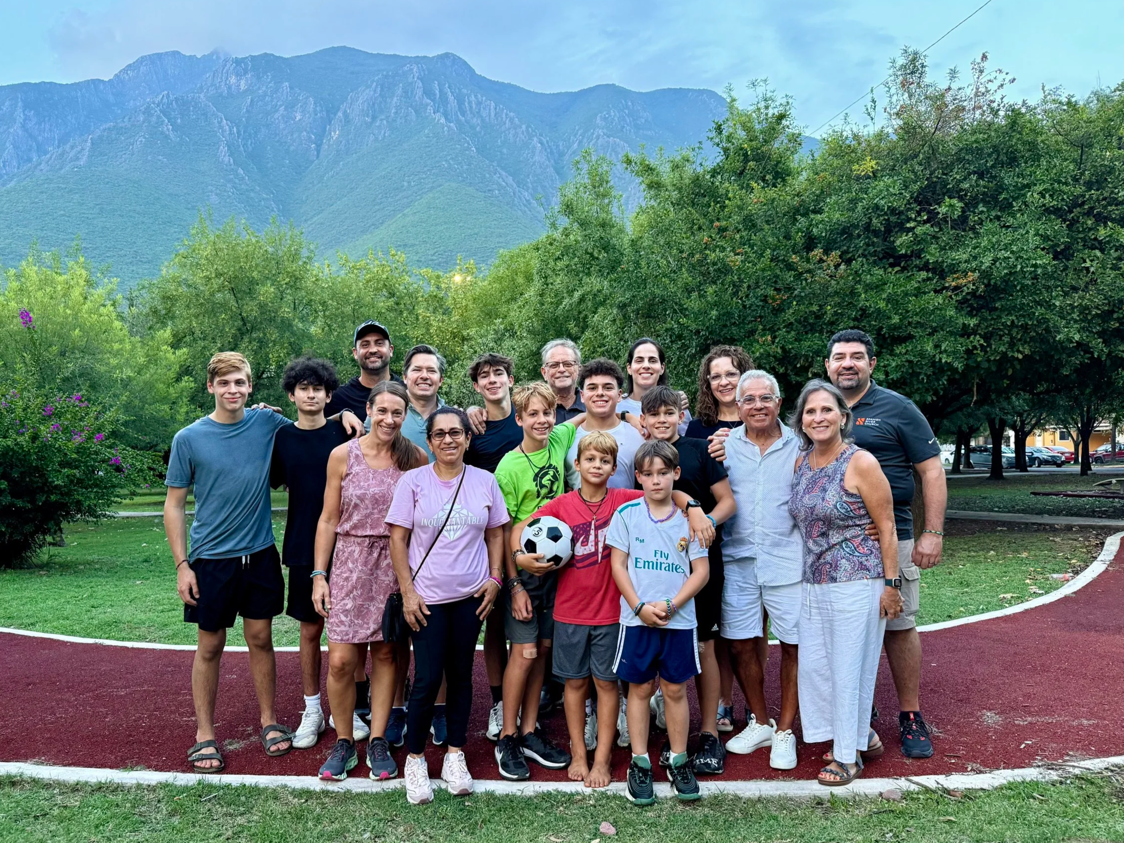 A group of people, including children, teenagers, and adults, standing outdoors on a track field with trees and mountains in the background during daytime.