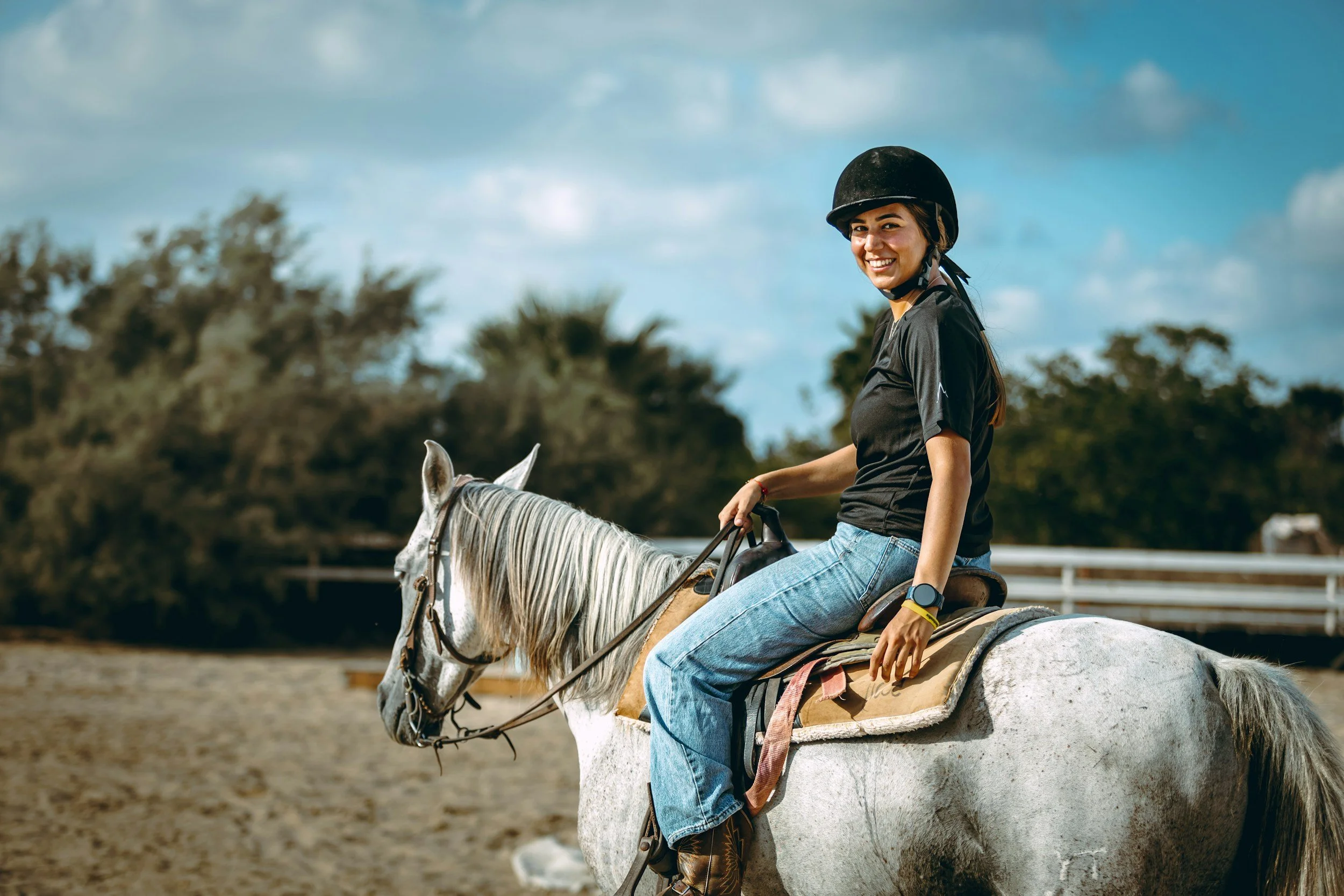 A woman riding a white horse outdoors during daytime, wearing a black helmet, black t-shirt, and jeans, smiling at the camera with trees and a cloudy sky in the background.