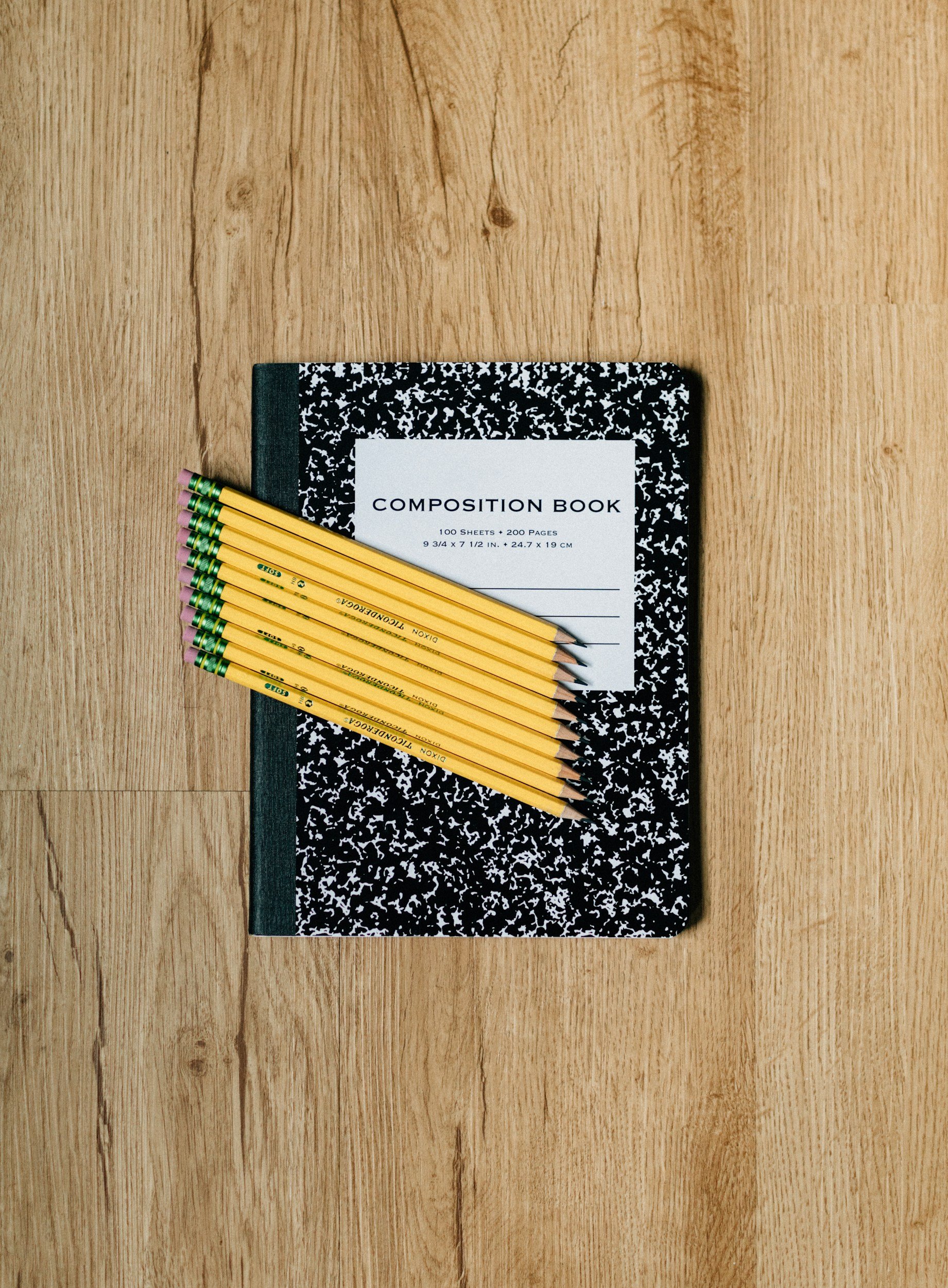 A composition notebook with a black and white marbled cover, labeled 'Composition Book' on a white sticker, next to several yellow pencils with pink erasers and green bands, all on a wooden surface.