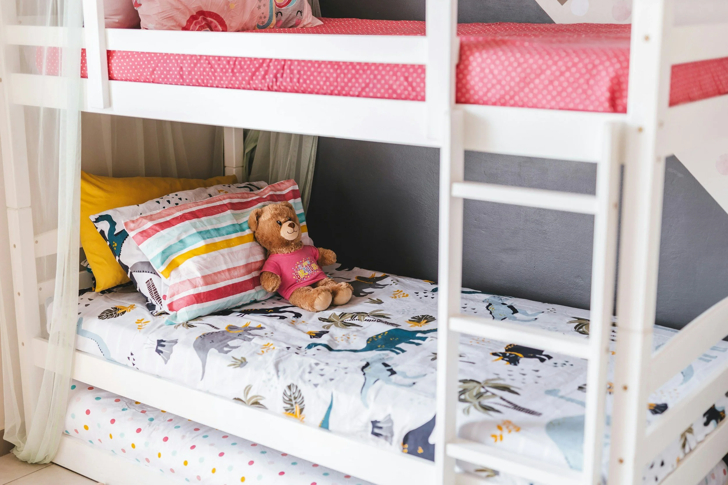 Bunk bed with pink polka dot bedding, a teddy bear wearing a pink shirt on the lower bed, and colorful pillows on the upper bed in a children's bedroom.