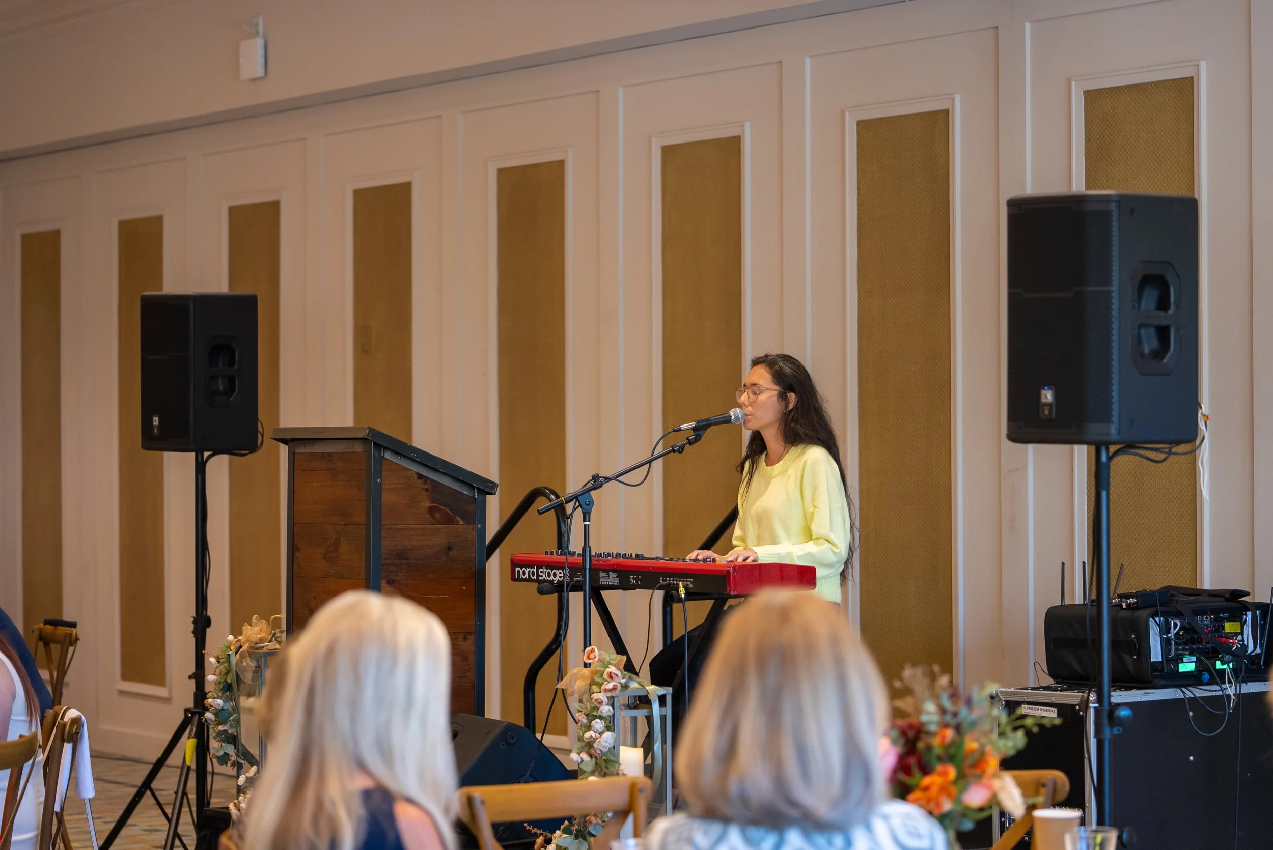 A young woman with glasses, wearing a yellow sweater, singing into a microphone while playing a red Nord Stage keyboard on a stage in a conference or event room. Two large black speakers are positioned on either side of her. The audience, with visibl