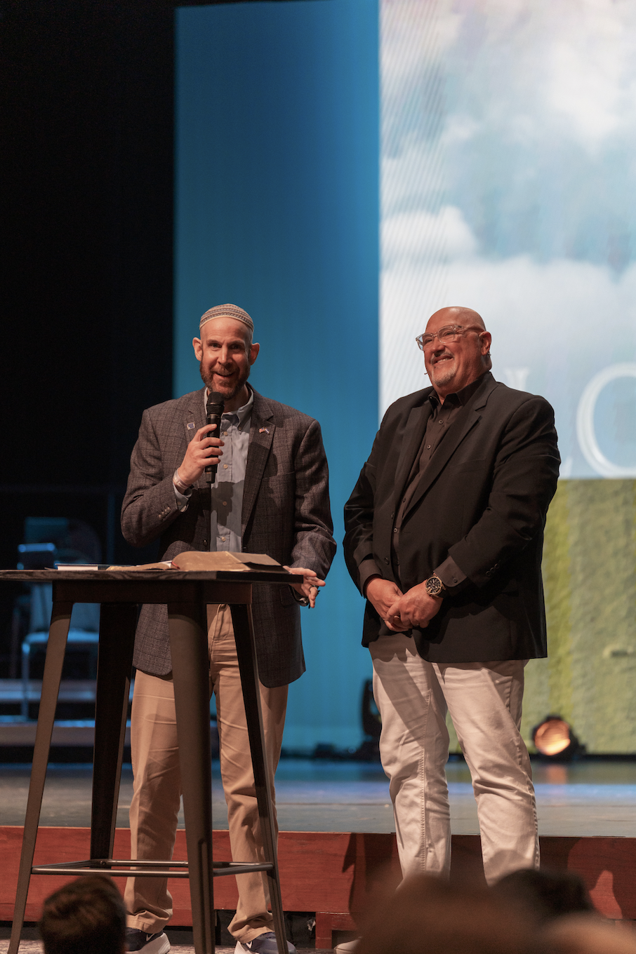 A smiling young man wearing an orange T-shirt with the text 'Spanish River Church' stands near a glass door that has similar signage. He has a black lanyard around his neck and a watch on his wrist.