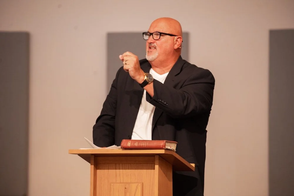 A man with glasses and a beard, wearing a black blazer over a white shirt, stands behind a wooden podium with a book on it. He appears to be speaking or presenting, with his right hand raised and a serious expression.