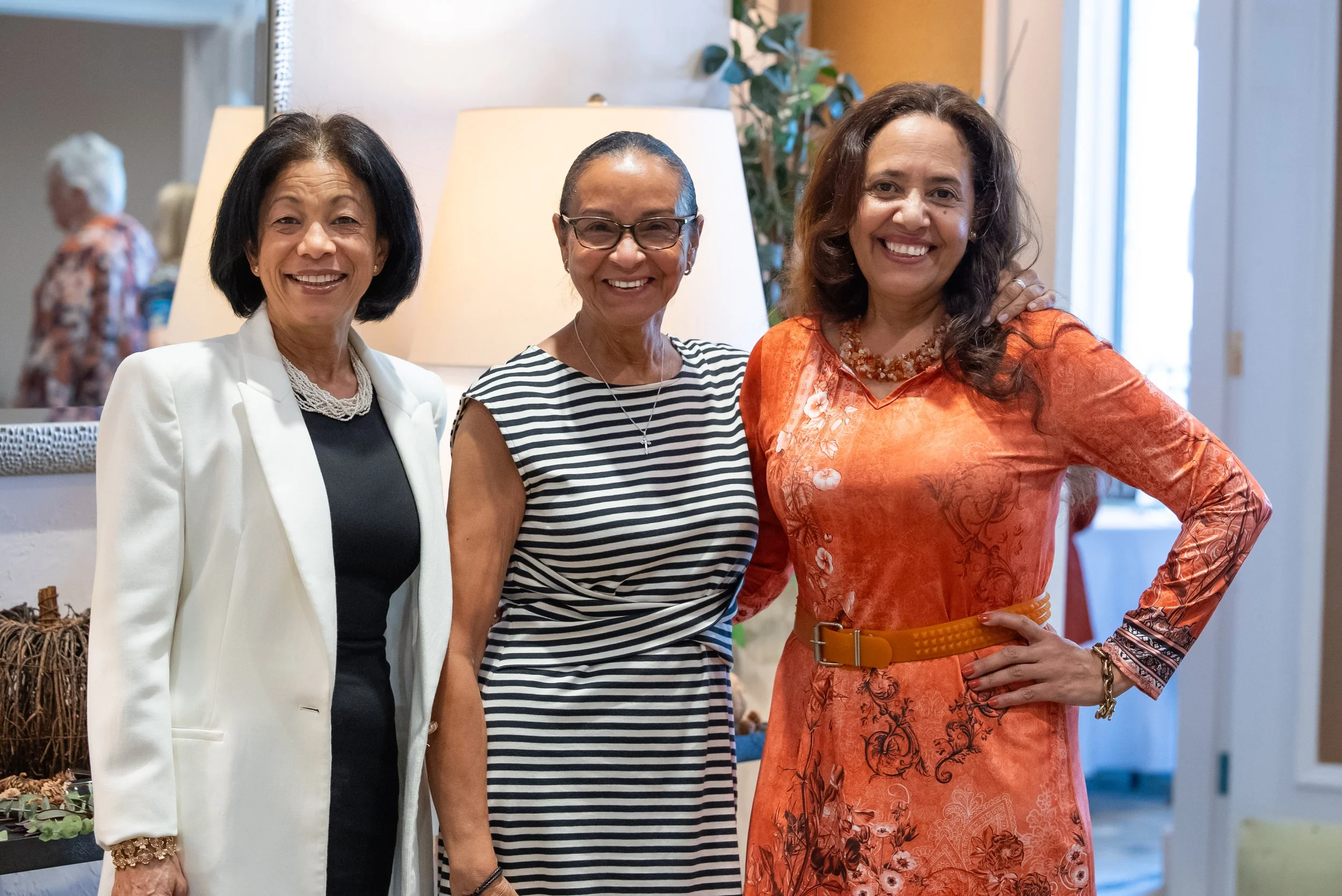 Three women smiling and standing close together indoors, with one woman's arm around the second woman's shoulder. The woman on the left has short black hair, wears a white blazer and pearl necklace. The woman in the middle has glasses, a striped top,