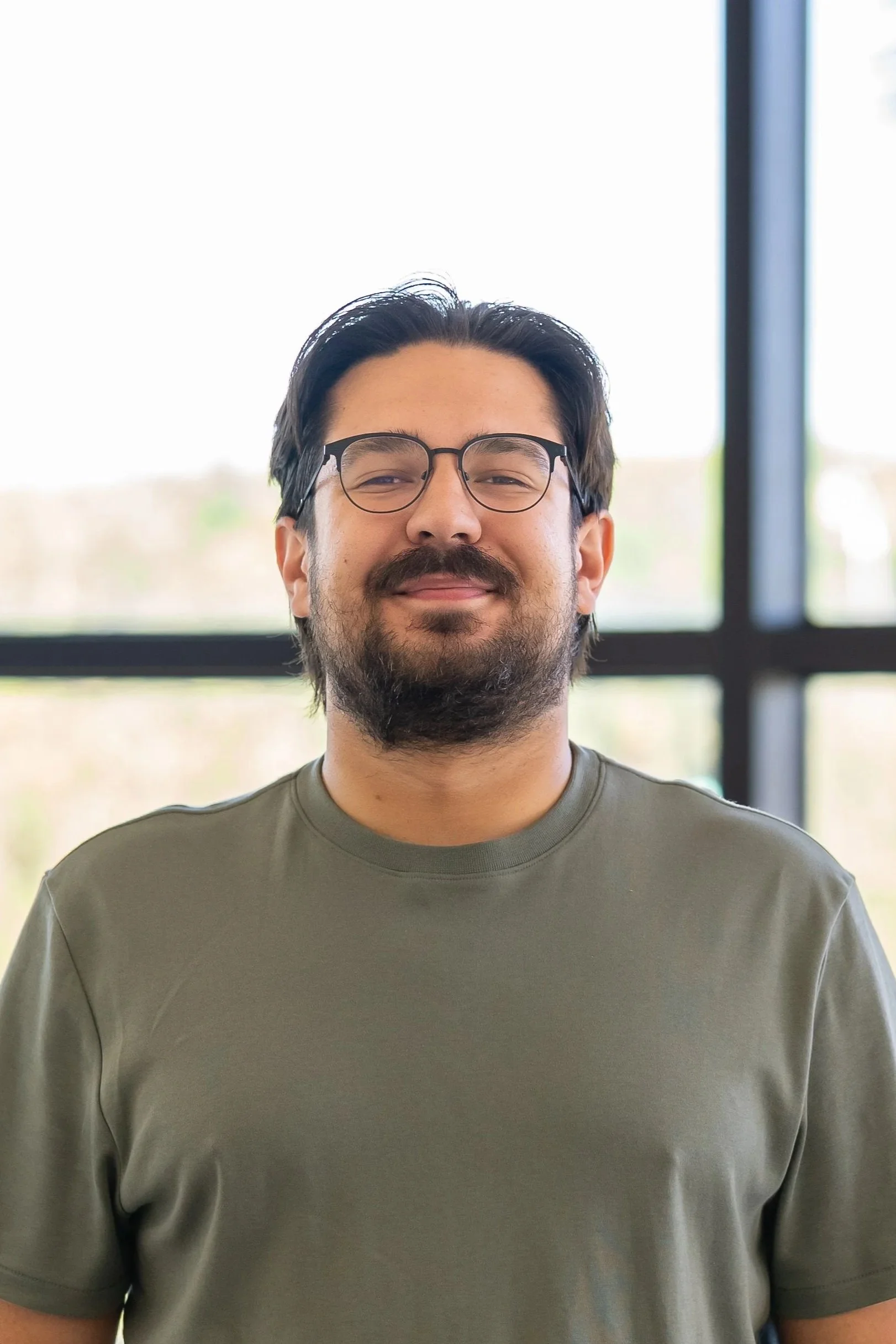 Close-up of a man with dark hair, glasses, and a beard, wearing a gray shirt, standing in front of large windows with a blurred outdoor background.