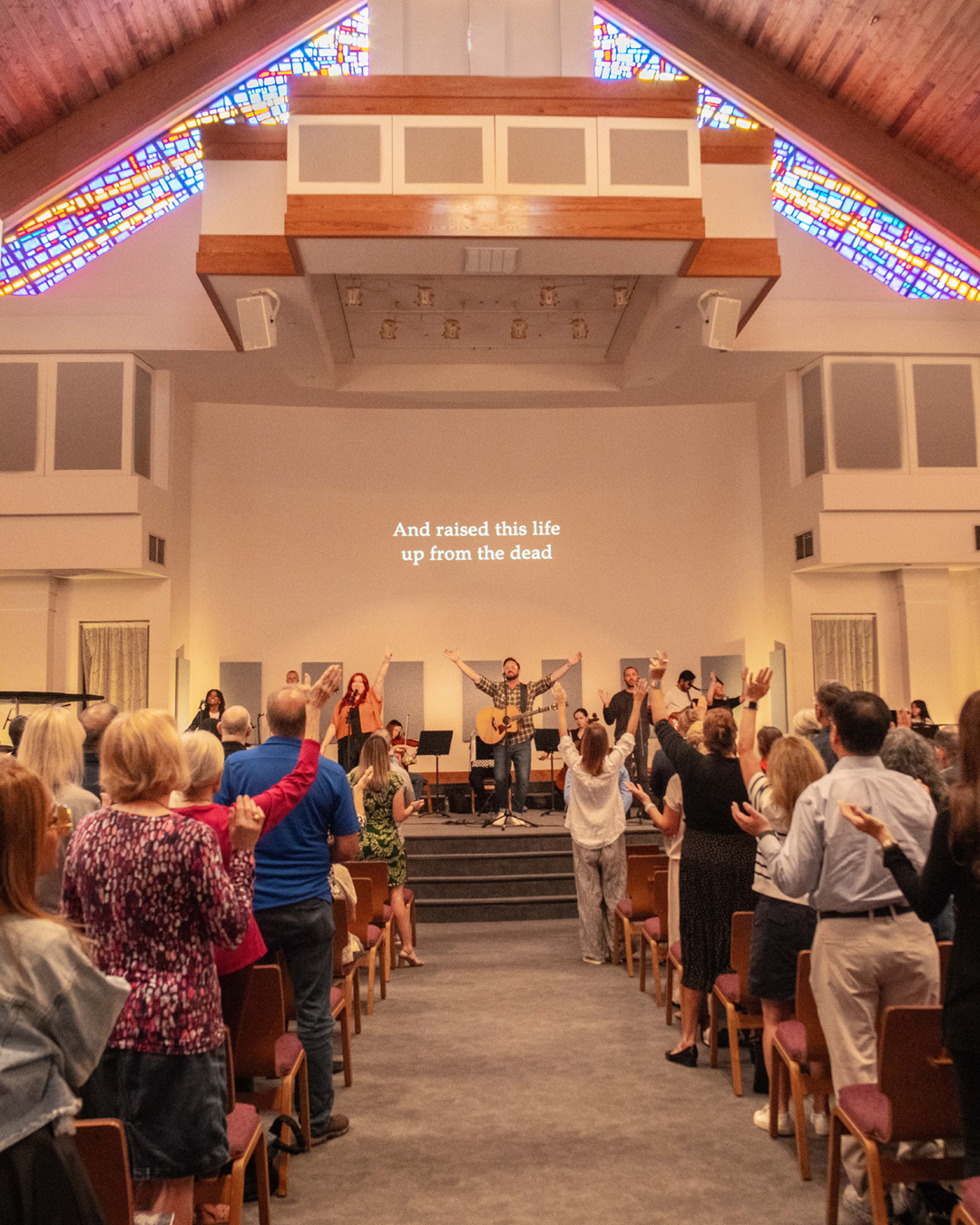 People singing and raising their hands in a church or concert setting, with a singer playing guitar on stage and lyrics projected on the wall that say, "And raised this life up from the dead."