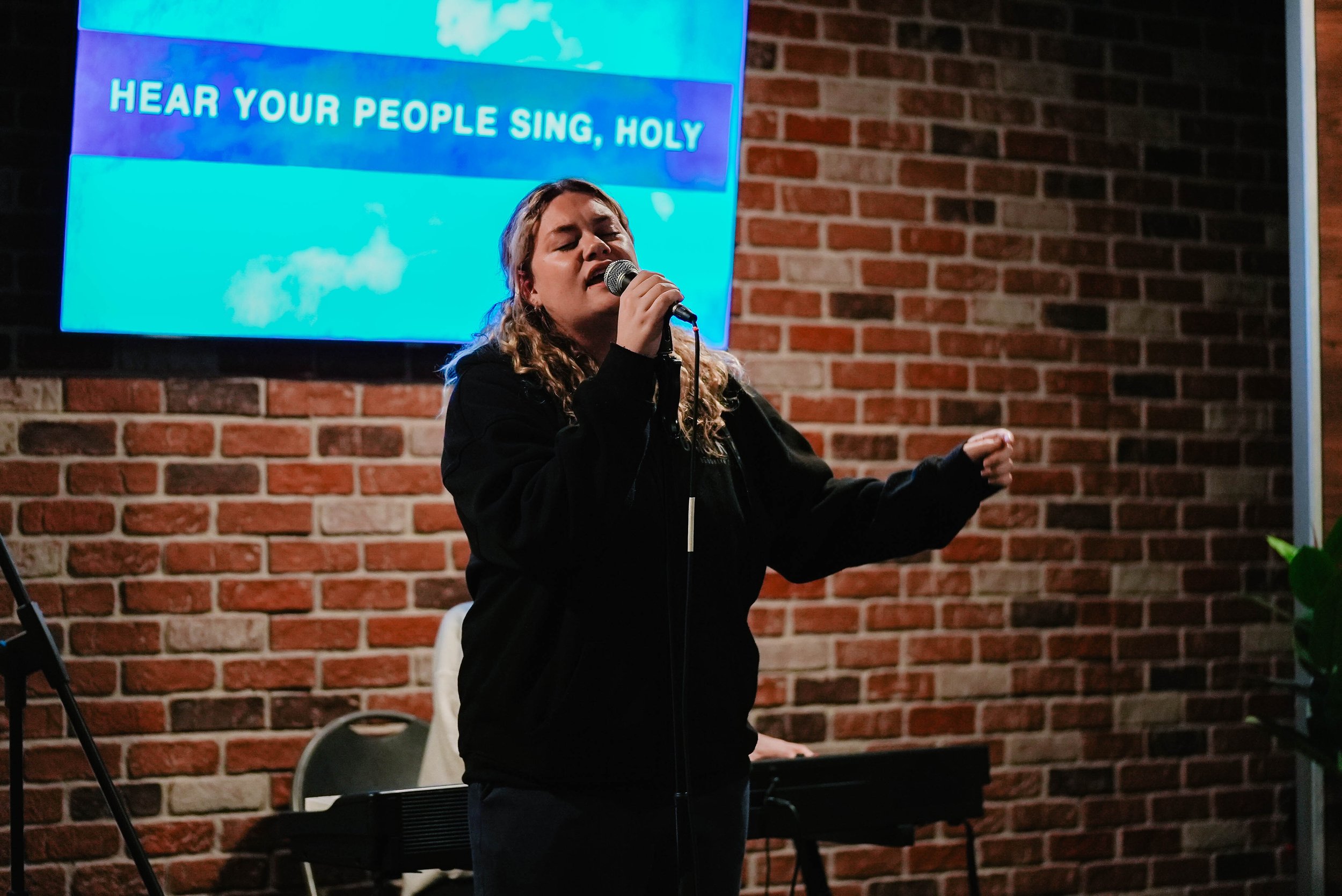 A woman singing into a microphone with closed eyes, standing in front of a brick wall, with a large screen displaying the words "HEAR YOUR PEOPLE SING, HOLY" behind her.