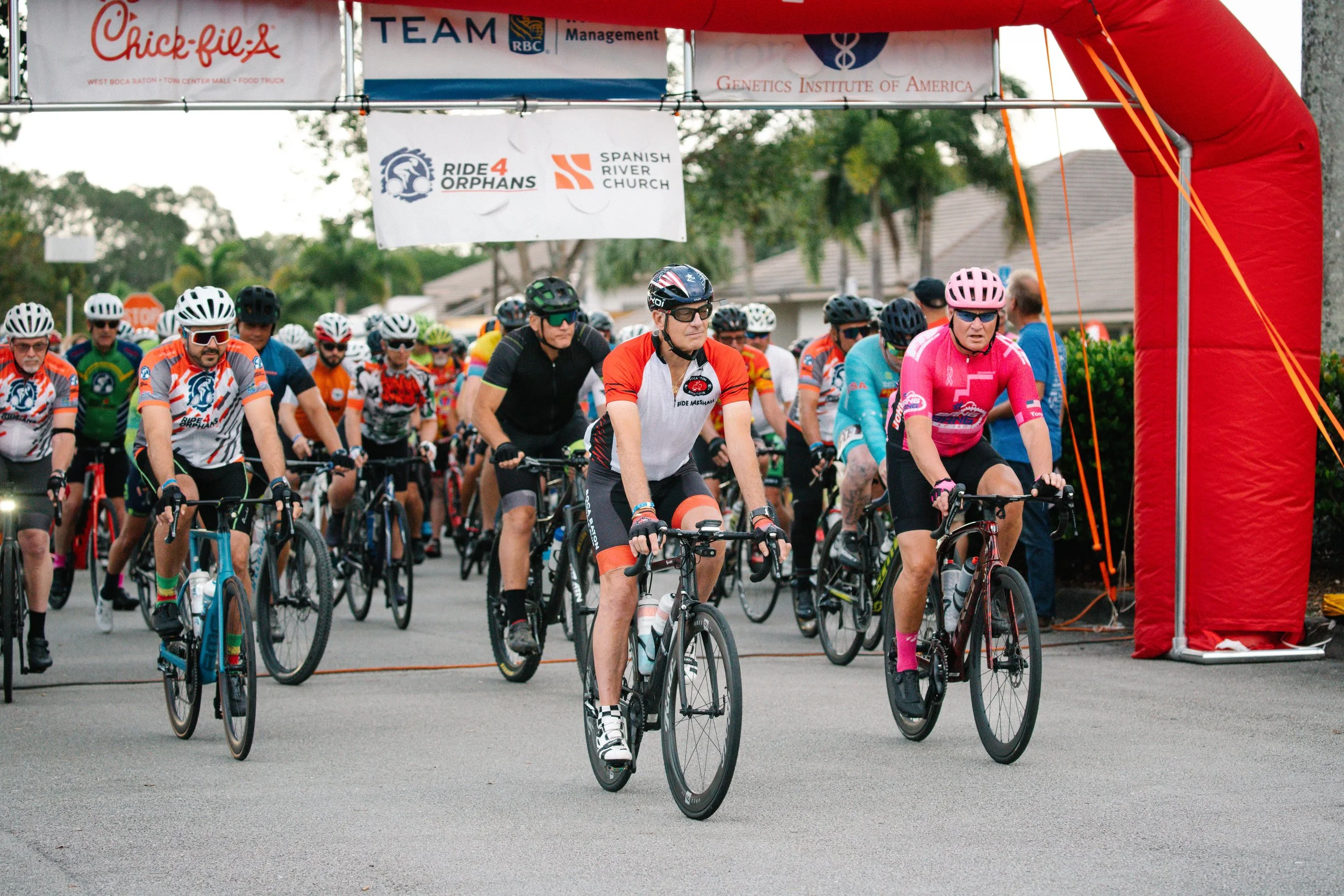 Cyclists at the start of a race under a red inflatable arch with sponsor banners, including Chick-fil-A and Genetics Institute of America, in a suburban neighborhood.