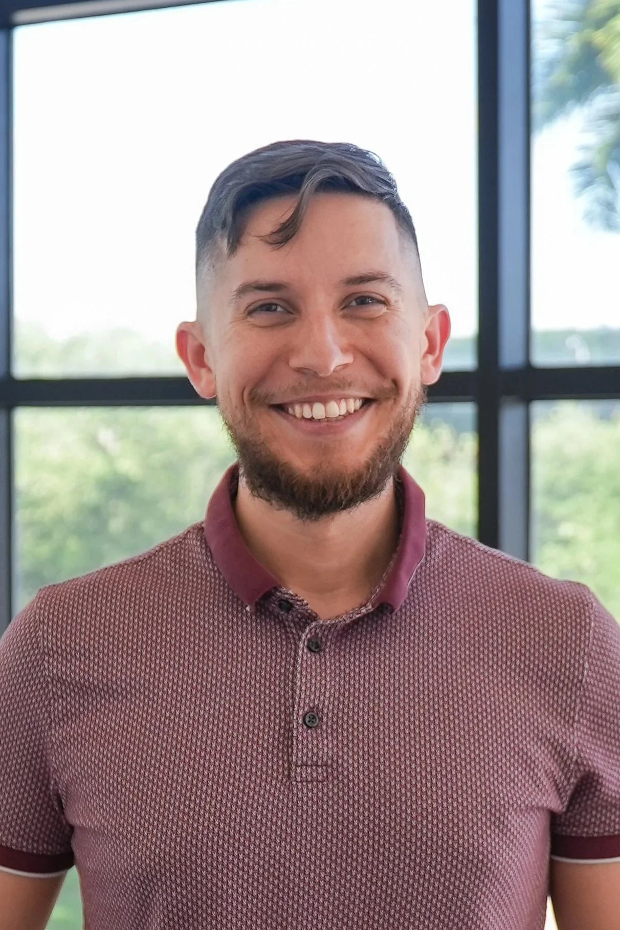 A smiling man with short dark hair and a beard, wearing a red patterned polo shirt, standing in front of large windows with a view of trees outside.