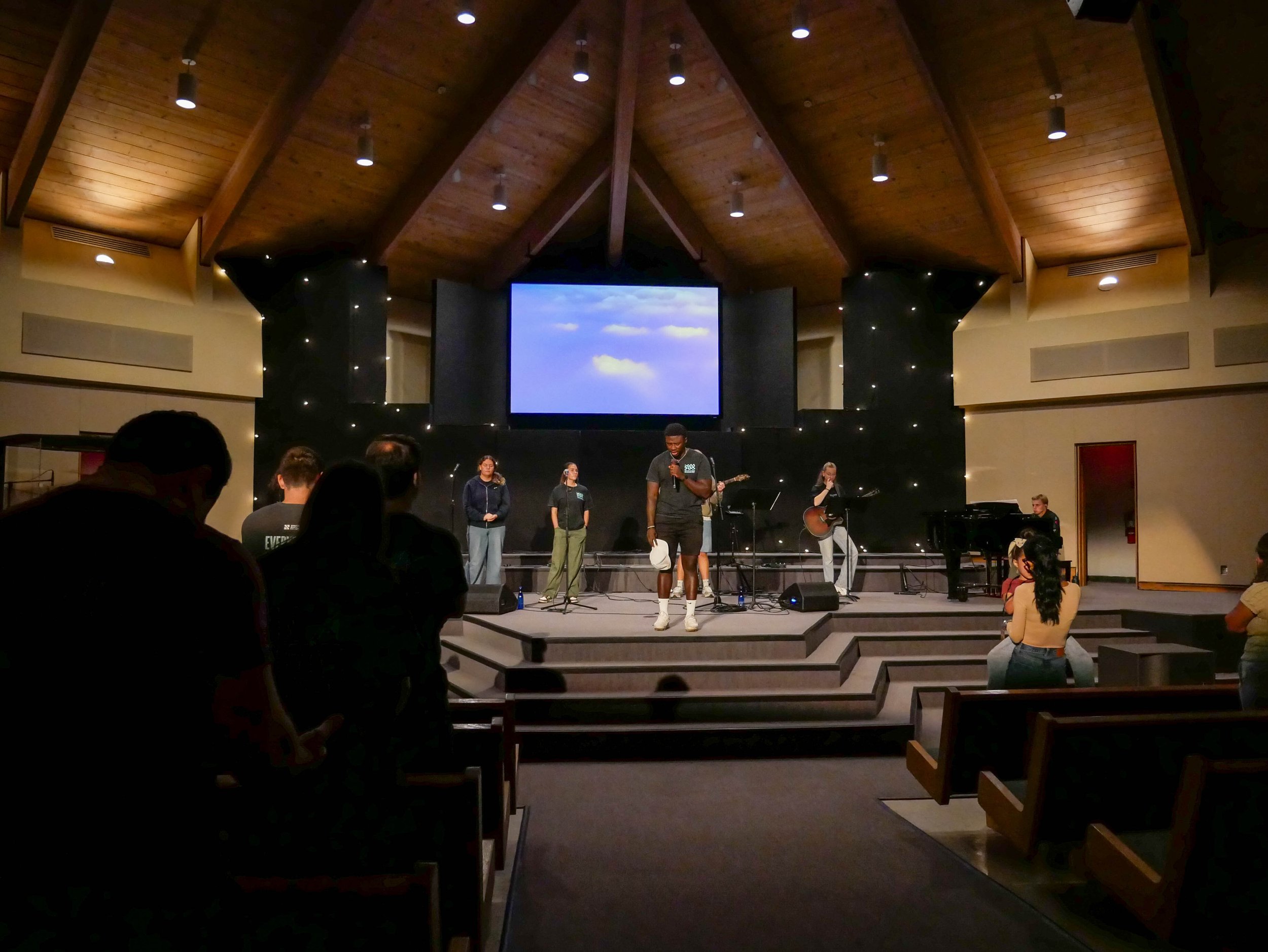 A group of people performing on a stage inside a church or auditorium. The stage has a large screen displaying a blue sky with clouds, and the ceiling above is made of wood with hanging lights. Some audience members are seated, facing the stage, and a few people are singing into microphones.