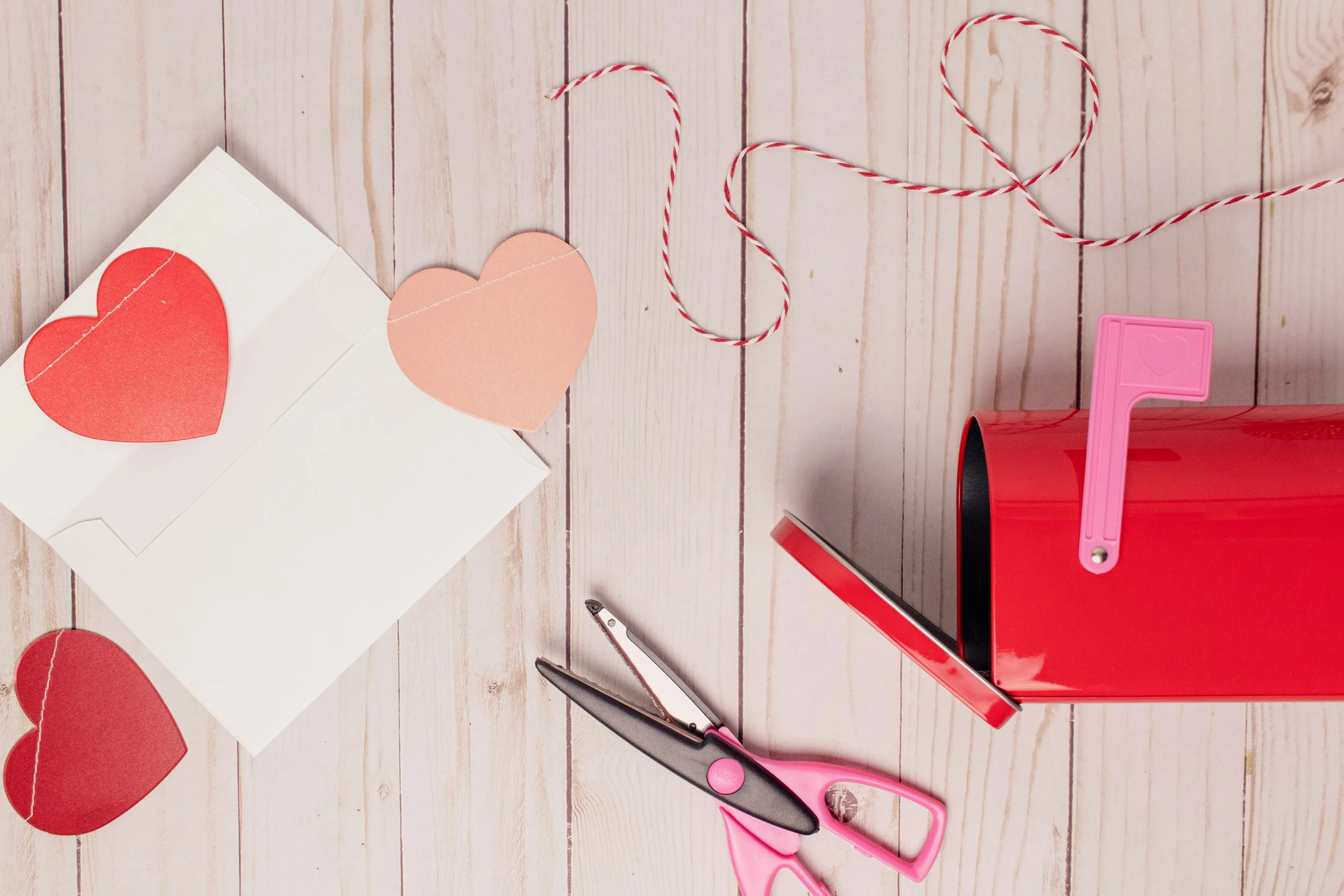 Pink scissors, a red gift box, a pink heart-shaped paper cutter, and heart-shaped paper cutouts on white paper with red and white twine on a light wooden surface.
