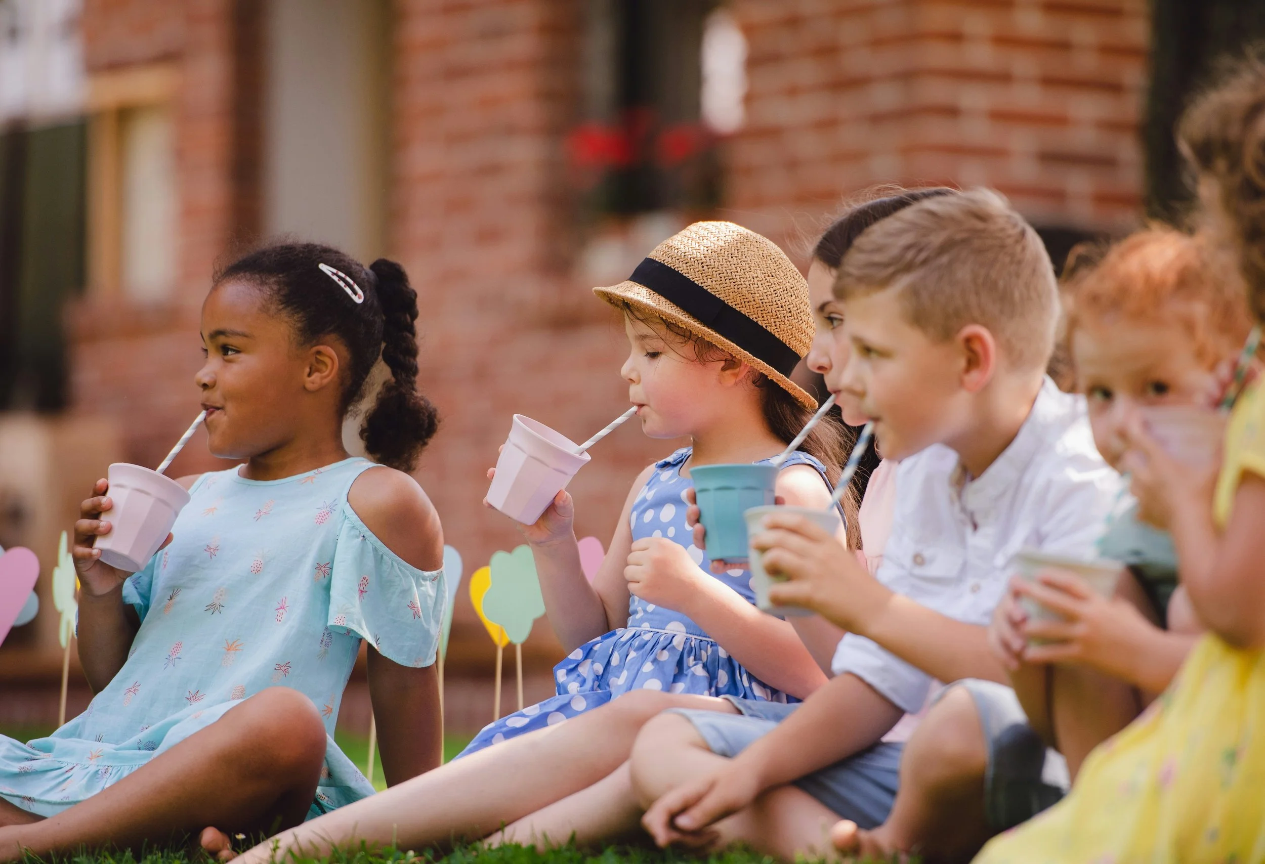 A group of children sitting outdoors on the grass, holding cups with straws, wearing colorful summer clothes, and a brick wall in the background.