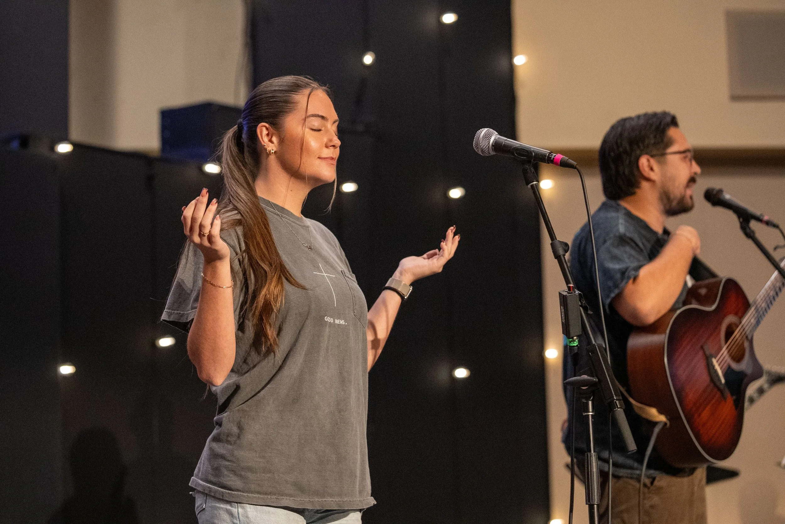 A young woman with long brown hair stands with eyes closed and hands raised, appearing to meditate or pray, while a man nearby plays guitar and sings into a microphone on stage.