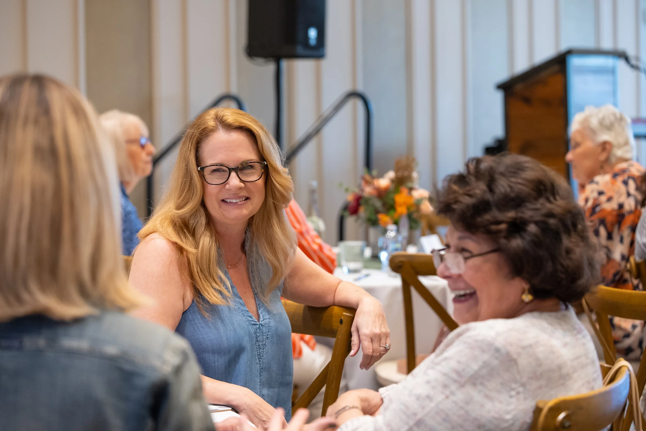 Group of women sitting at a table, engaging in conversation at a social gathering, with a woman with red hair and glasses smiling in the center.