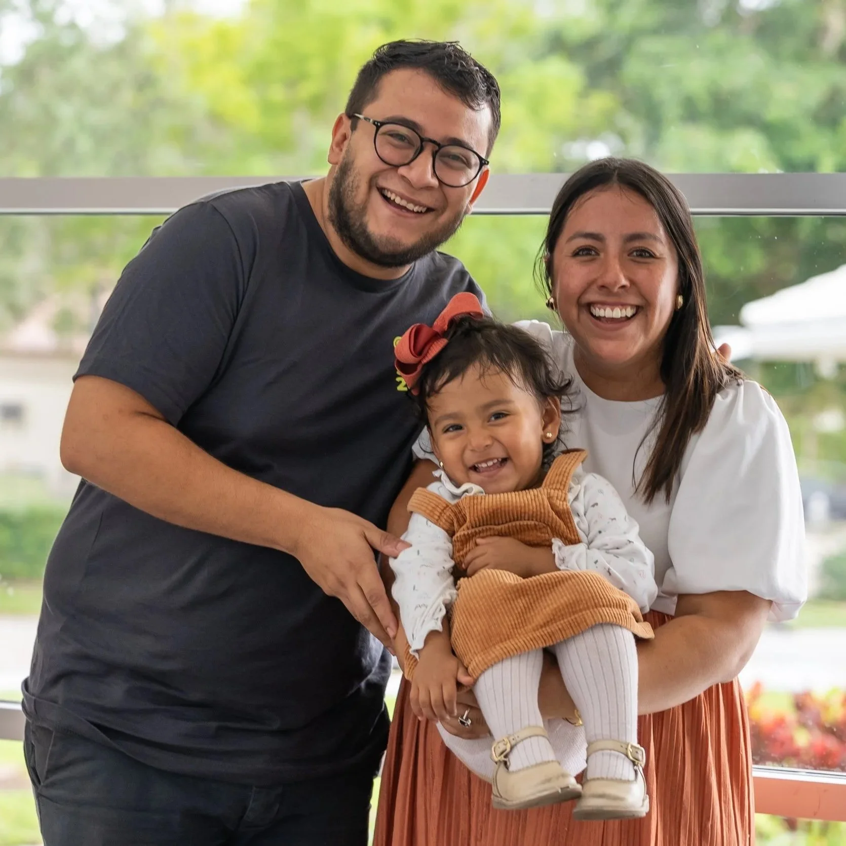 A happy family of three: a man, a woman, and a young girl, smiling and posing for a photo inside a building with a large window showing trees outside.
