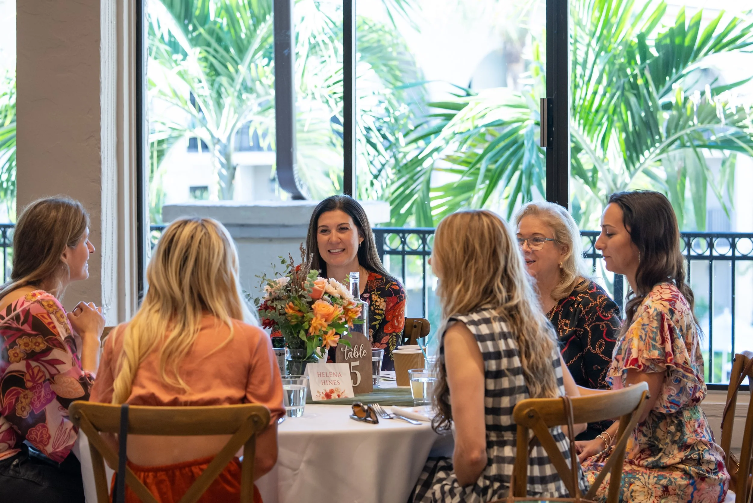 Group of women seated around a dining table engaged in conversation, with a woman in the center smiling. The table has a floral centerpiece and is set with glasses and plates. Bright natural light streams in through large windows with a view of green foliage outside.