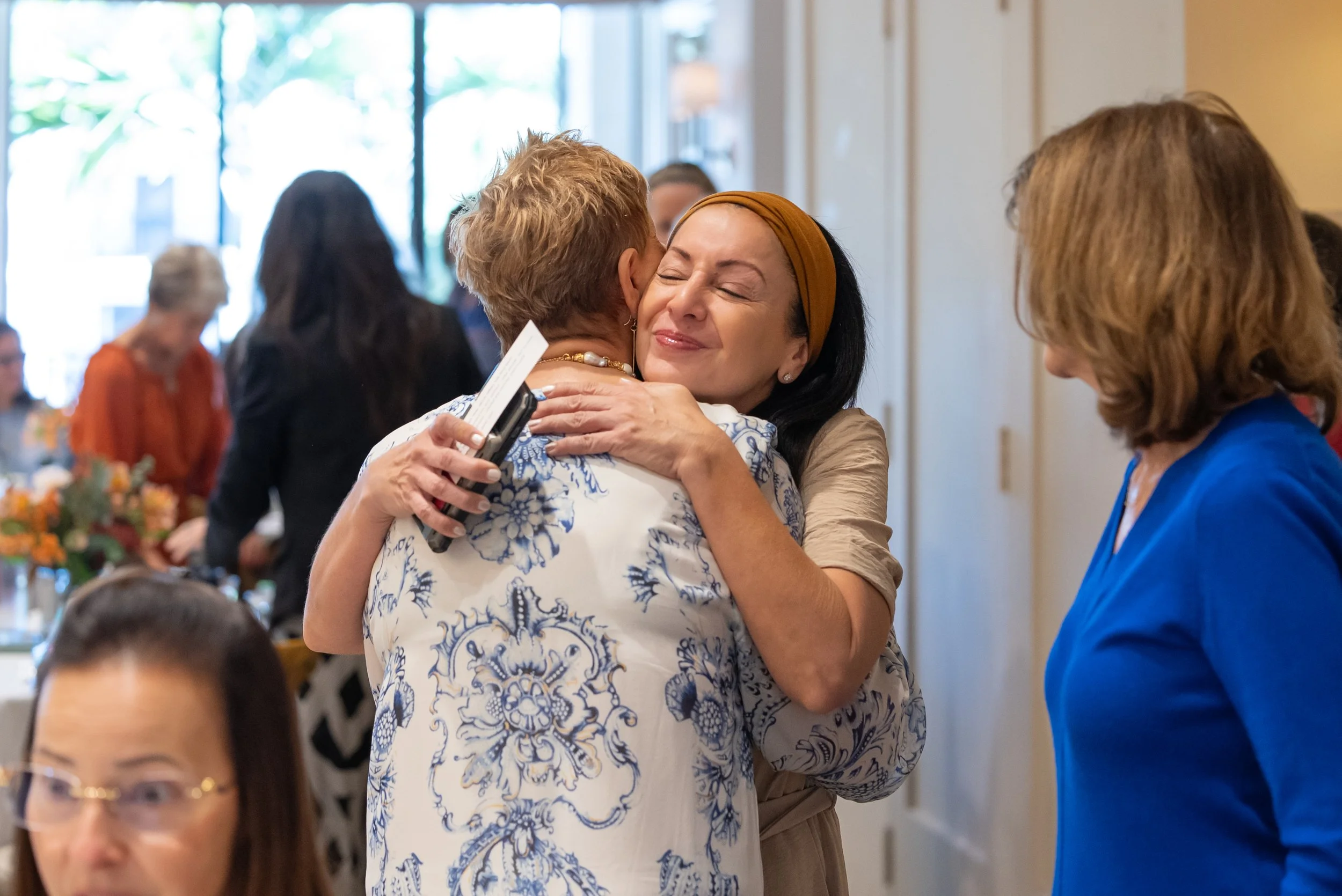 Two women hugging at a social gathering with other attendees in the background.