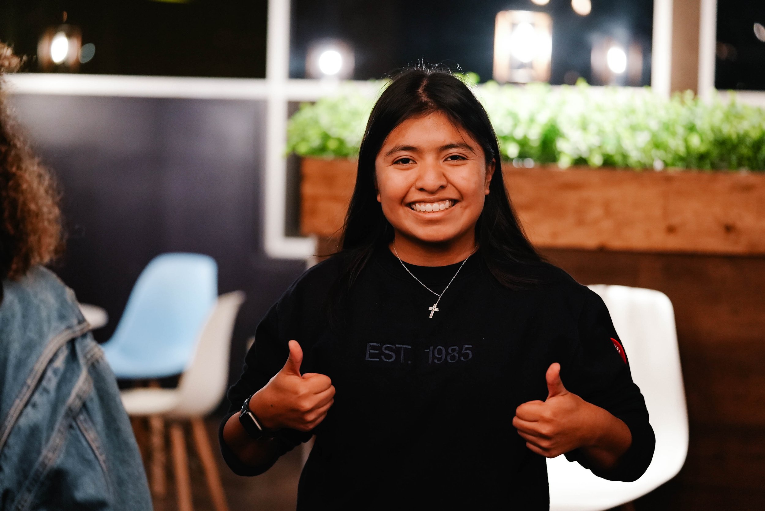 Smiling woman giving thumbs up in a modern indoor setting with plants in the background.