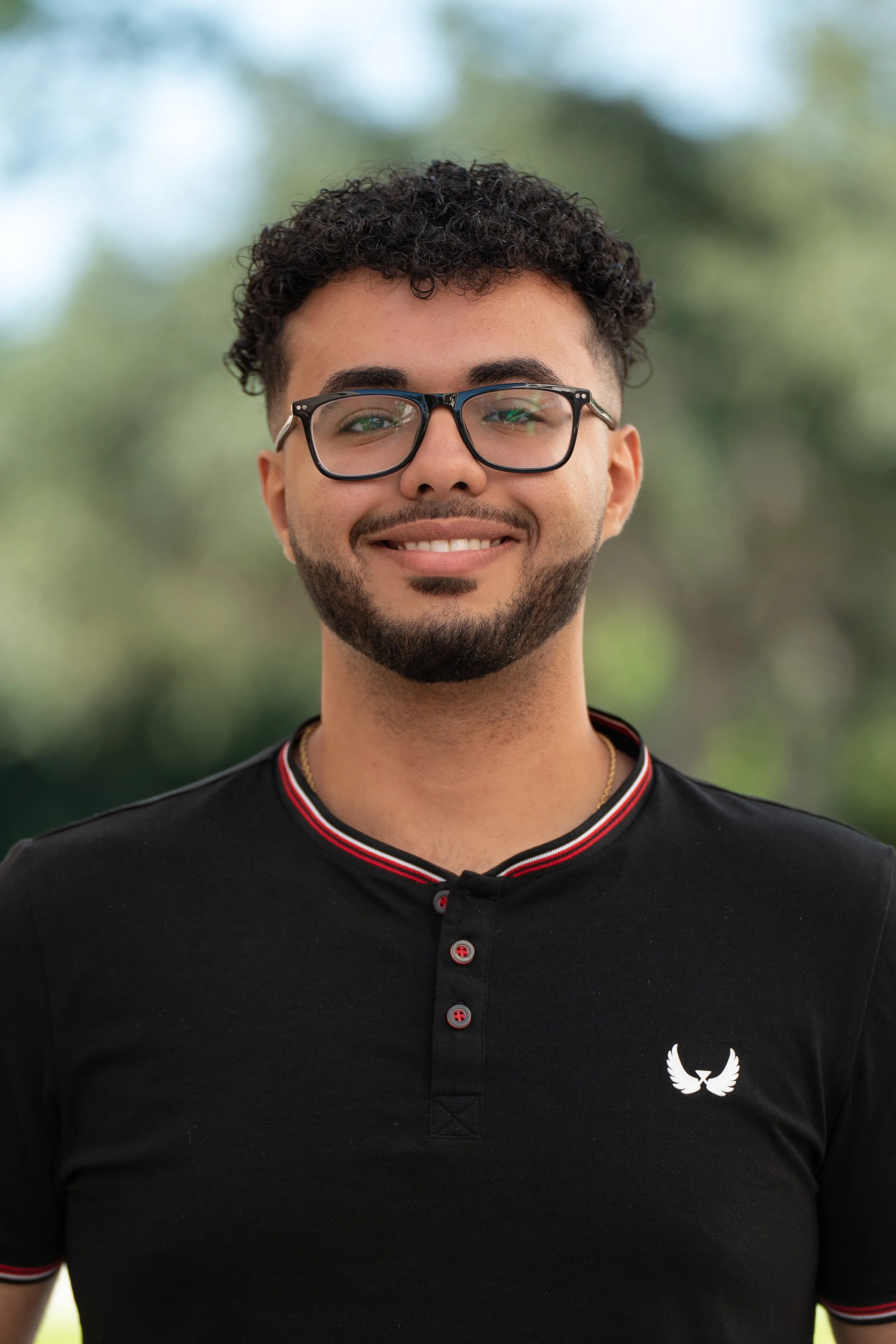 A young man with curly dark hair, glasses, a beard, and a mustache, smiling, wearing a beige shirt and a gold cross necklace, standing in front of a window with natural light.
