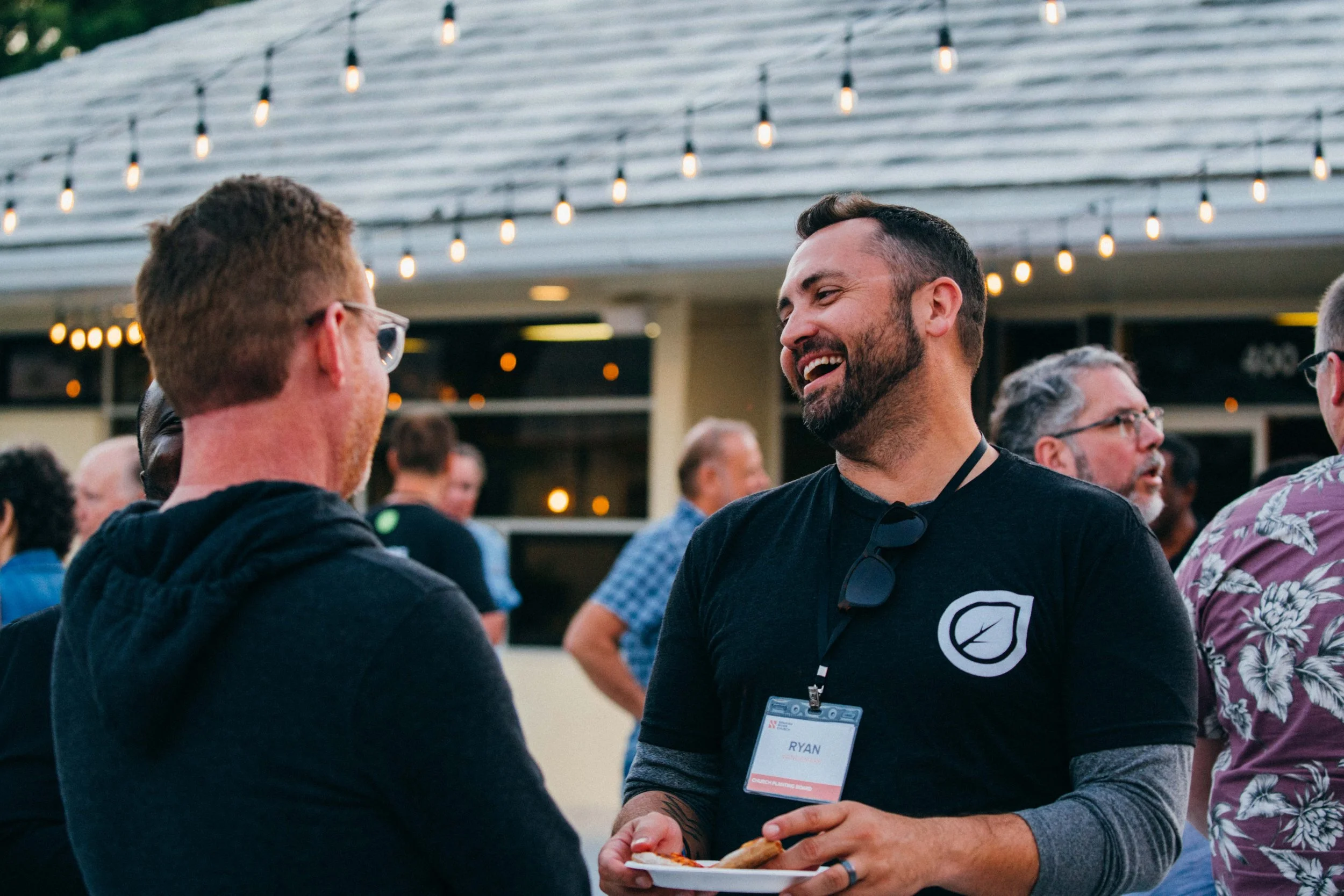 Two men are talking and laughing at an outdoor event during the evening, with string lights overhead. One man has a beard, is wearing a black shirt with a logo, and has sunglasses hung around his neck. The other man has glasses and reddish hair. Other people are visible in the background.