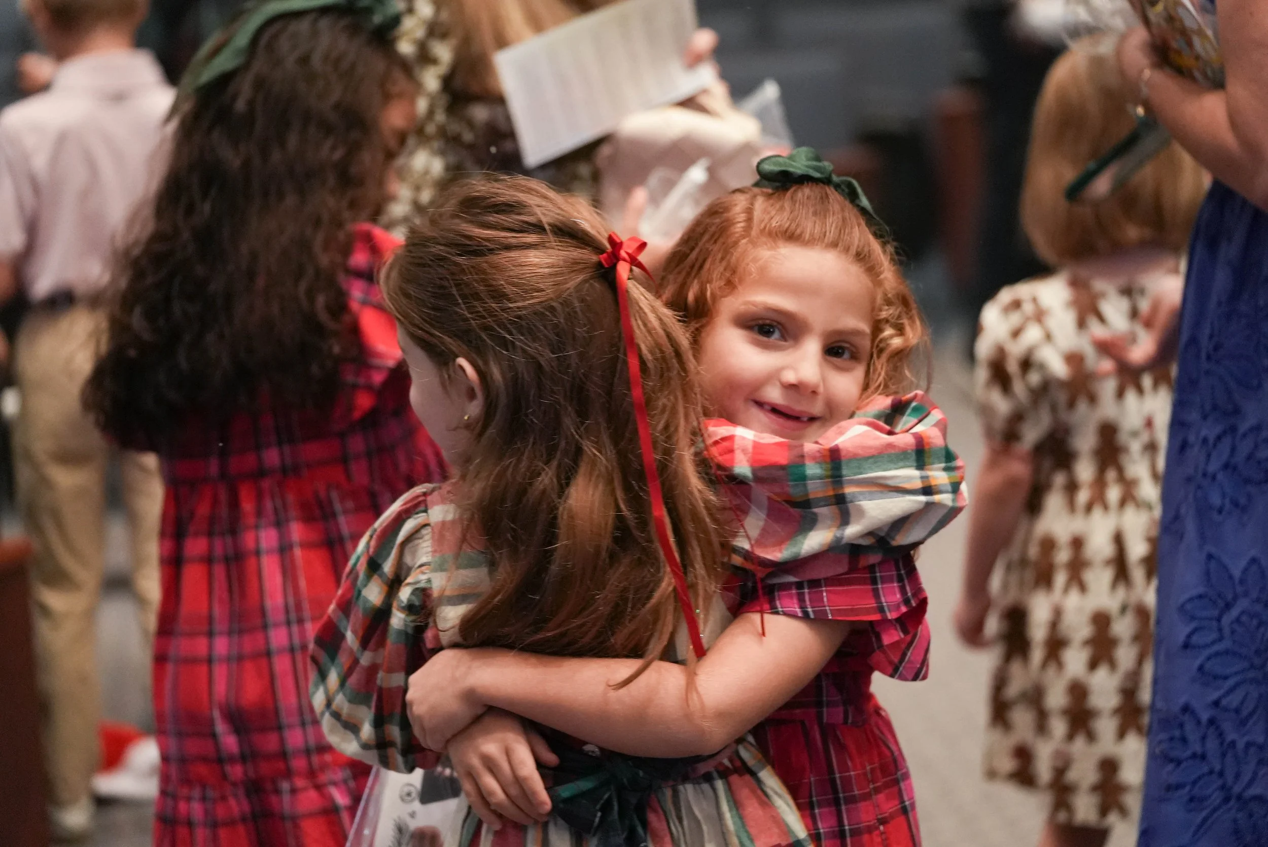 Two young girls wearing plaid dresses hugging each other, smiling at the camera, in a festive gathering.