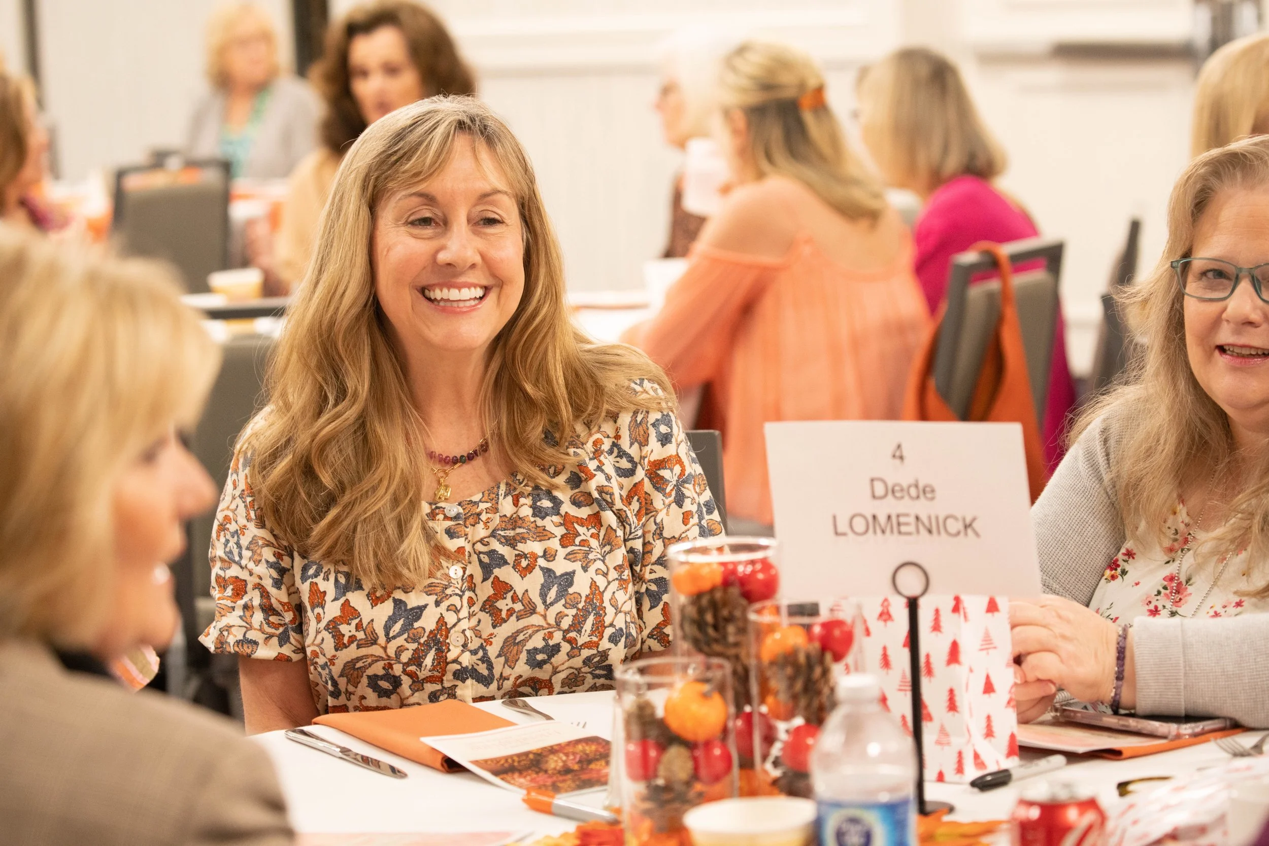 A smiling woman with long wavy hair sitting at a table during a group event called Friendsgiving. The table has festive decorations and a bottle of water, with other women sitting around.