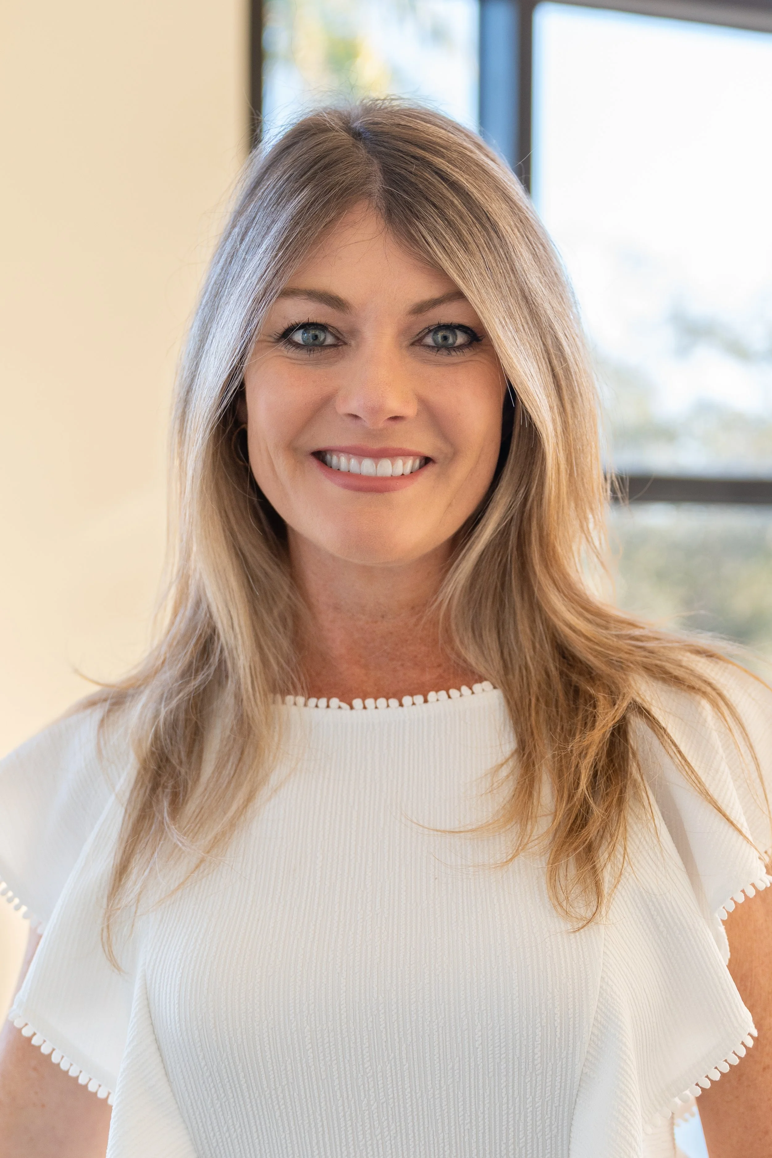 A woman with blonde hair and blue eyes smiling outdoors with blurred greenery in the background.