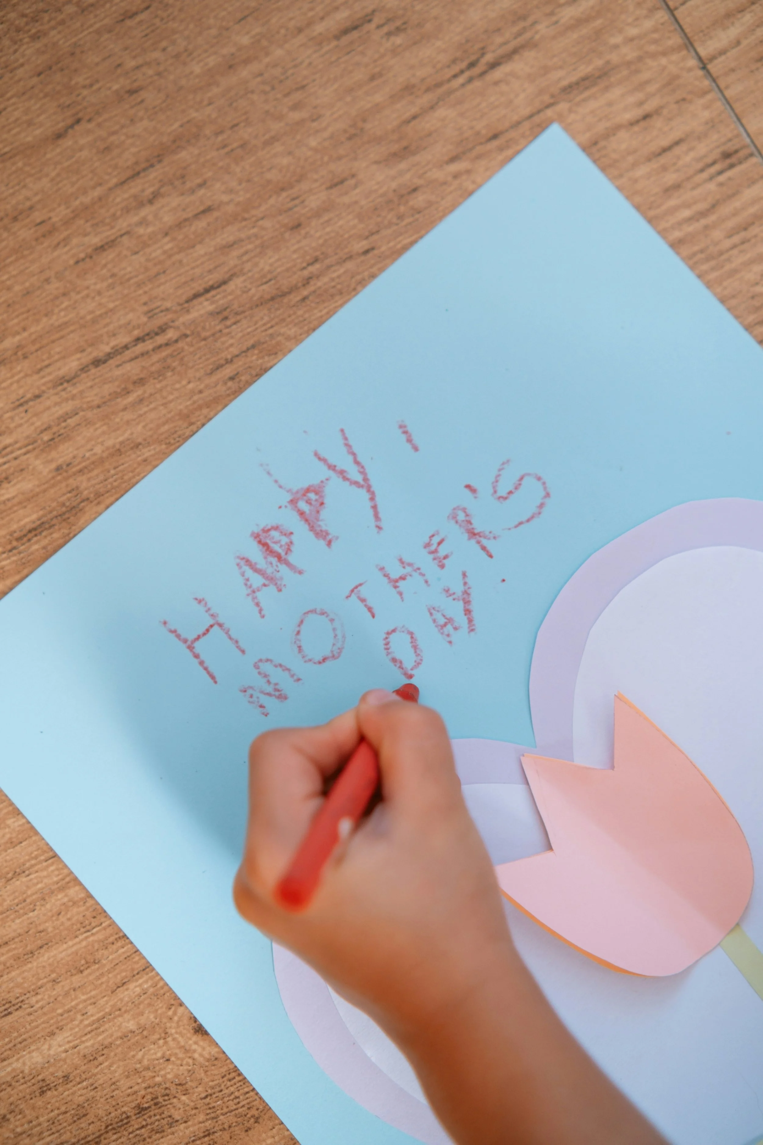 Child writing on a blue paper with red crayon, creating a greeting card with a pink paper flower attached to the card.