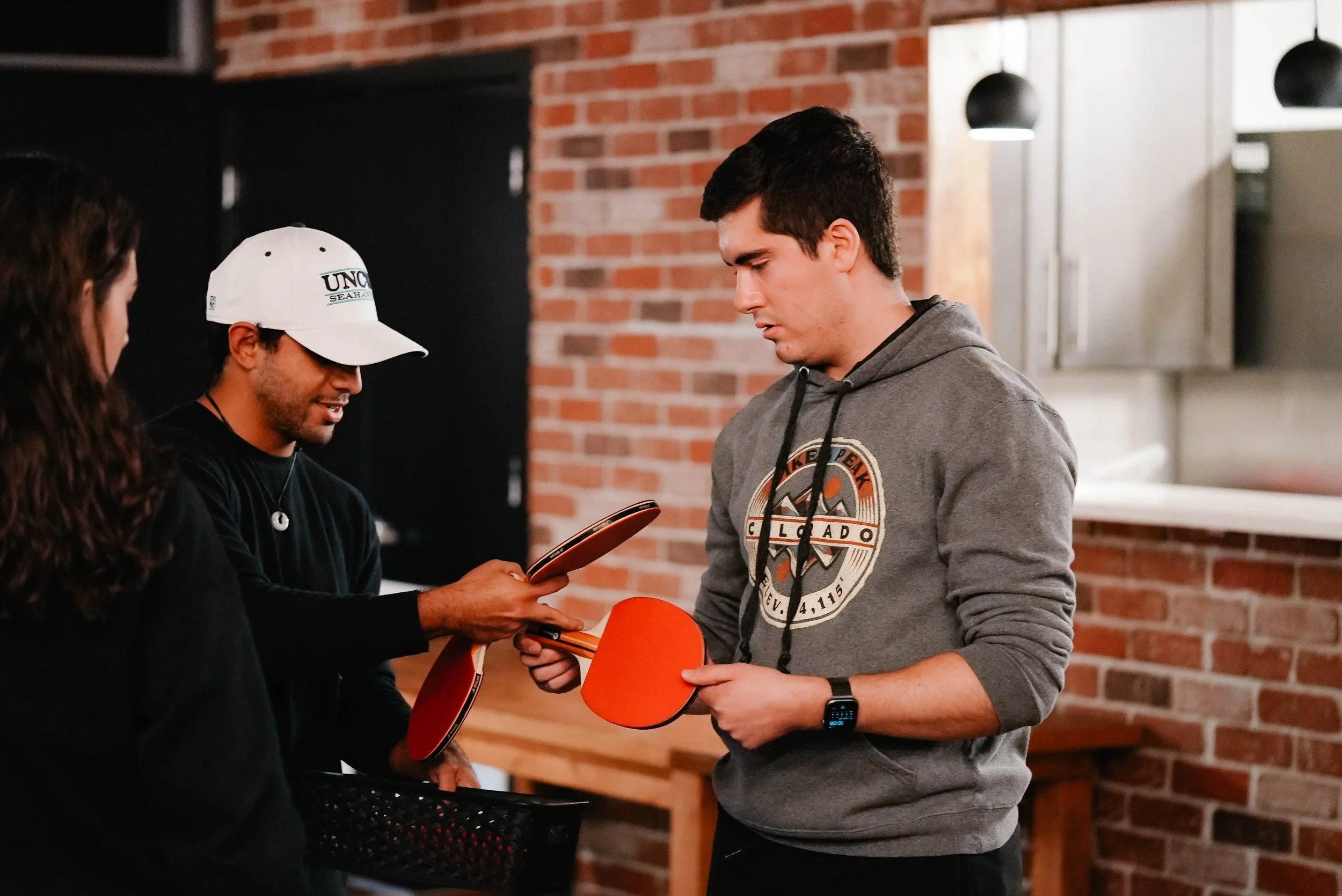 Two young men are holding orange paddle tennis rackets, and they appear to be in a conversation or exchanging items in an indoor setting with a brick wall background.