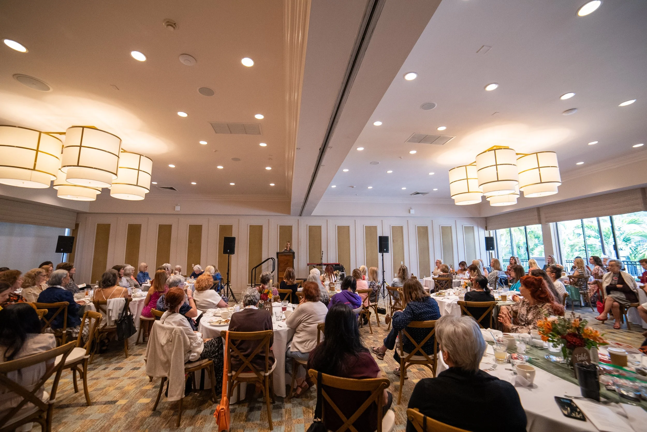 A large banquet hall filled with many women seated at round tables for a formal event, with a speaker at a podium on a stage at the front. The room has high ceilings, large hanging light fixtures, and windows letting in natural light.