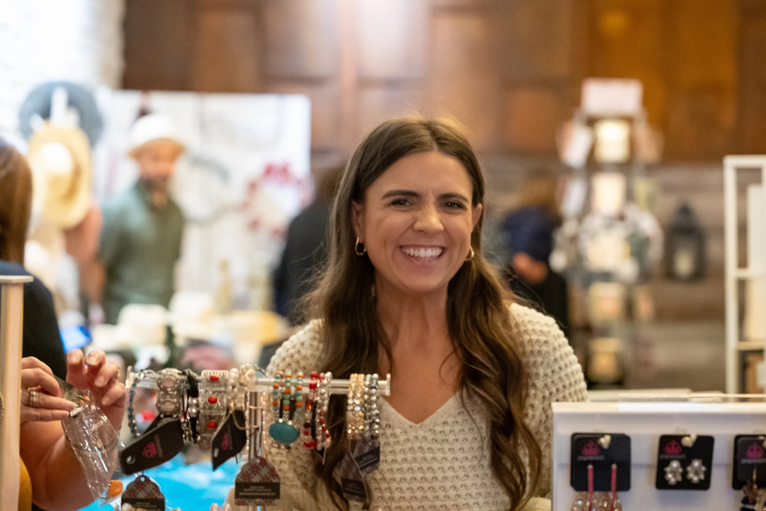 A woman with long brown hair smiling at a jewelry store counter displaying earrings and other jewelry items.