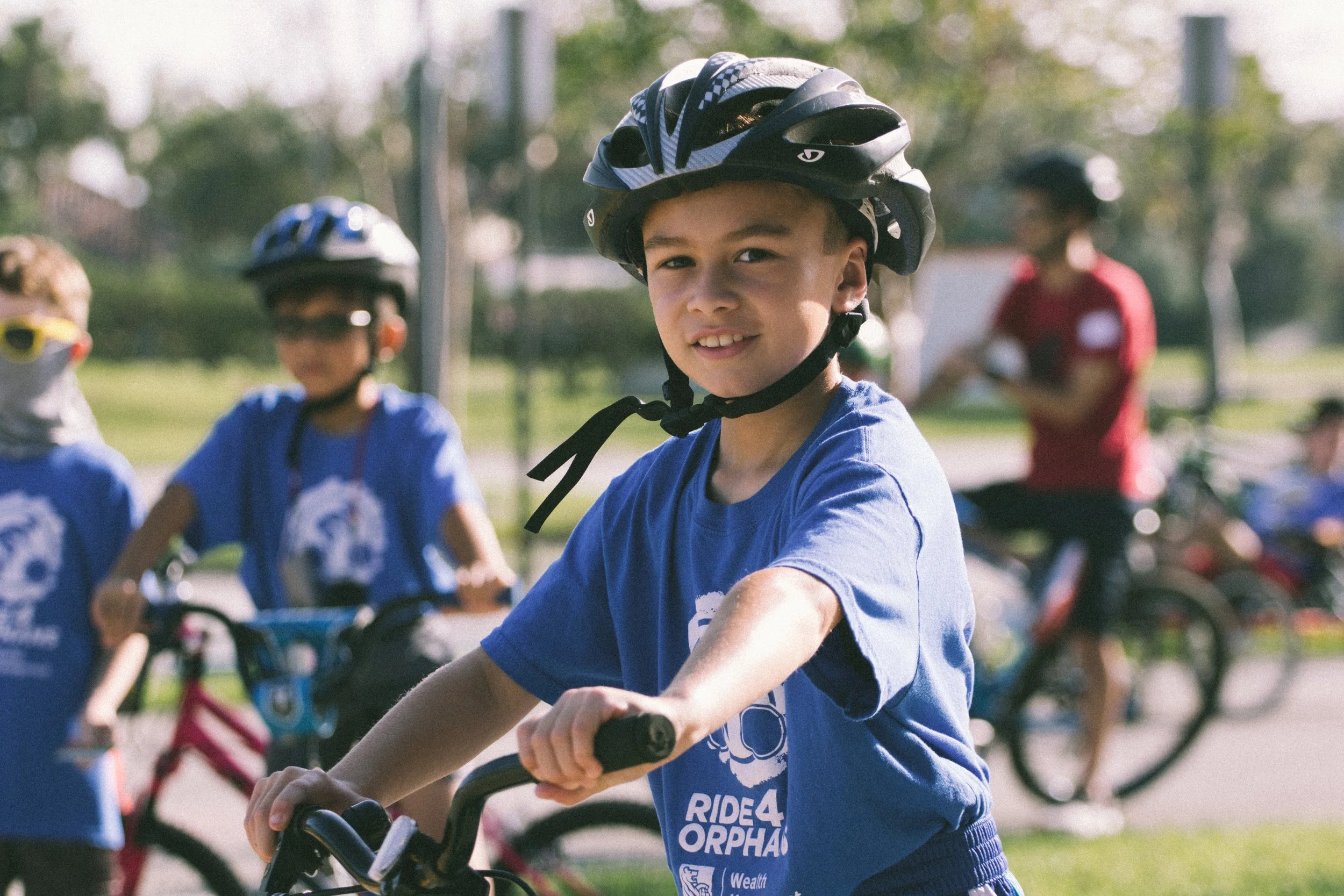 Cyclists at the start of a race under a red inflatable arch with sponsor banners, including Chick-fil-A and Genetics Institute of America, in a suburban neighborhood.