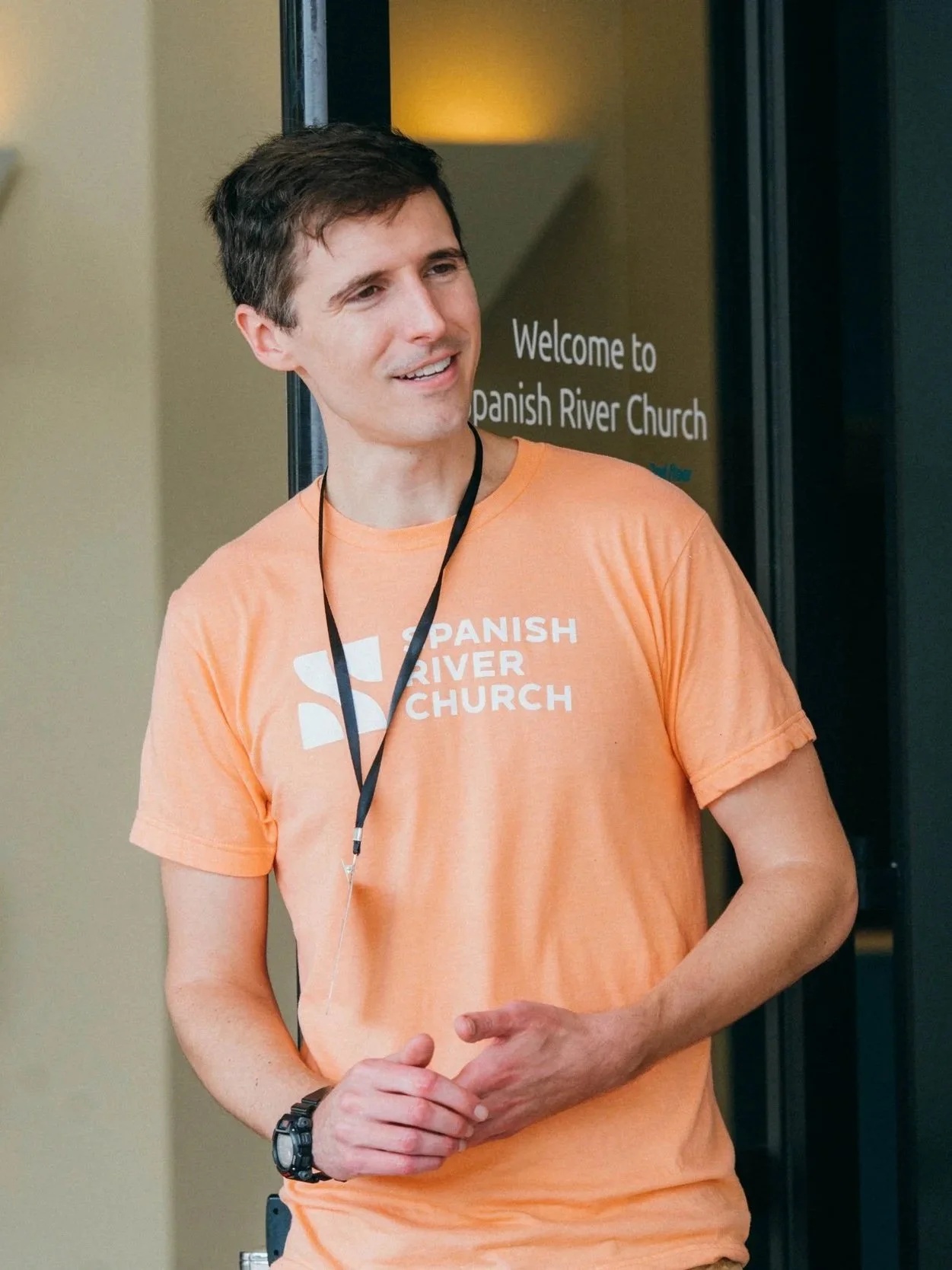 A smiling young man wearing an orange T-shirt with the text 'Spanish River Church' stands near a glass door that has similar signage. He has a black lanyard around his neck and a watch on his wrist.
