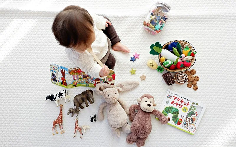 Young child sitting on white blanket surrounded by stuffed animals, toys, and books, with colorful craft supplies nearby.