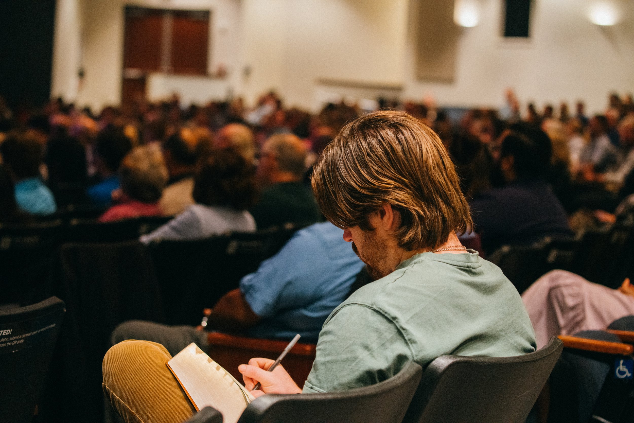 A man with brown hair reading a notebook and taking notes while seated in a crowded auditorium.