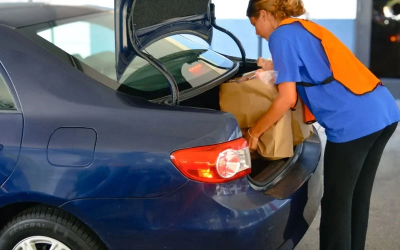A woman loading grocery bags into the trunk of a dark blue sedan at a gas station.