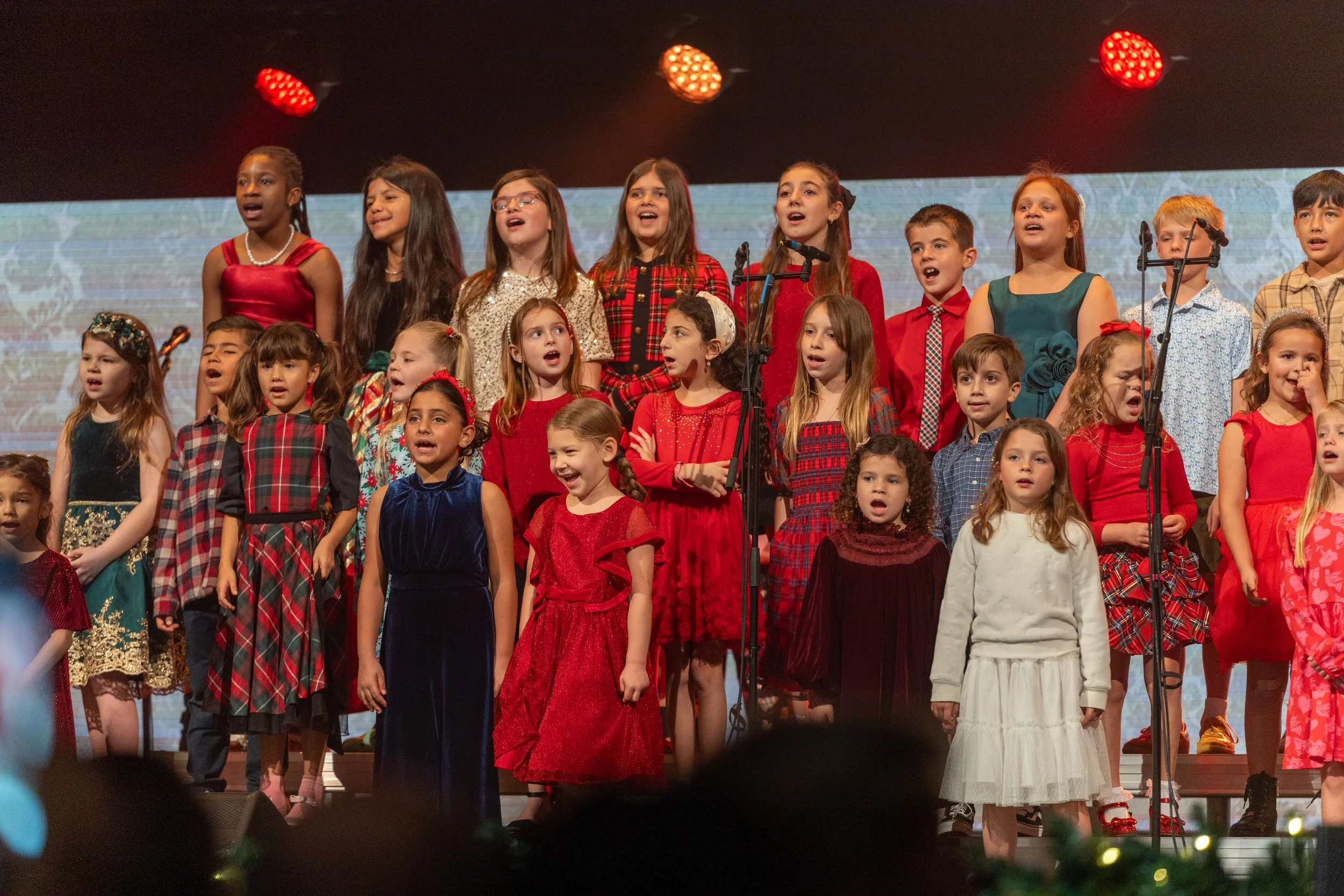 A group of children singing on stage during a holiday performance, dressed in festive red, green, and white attire.