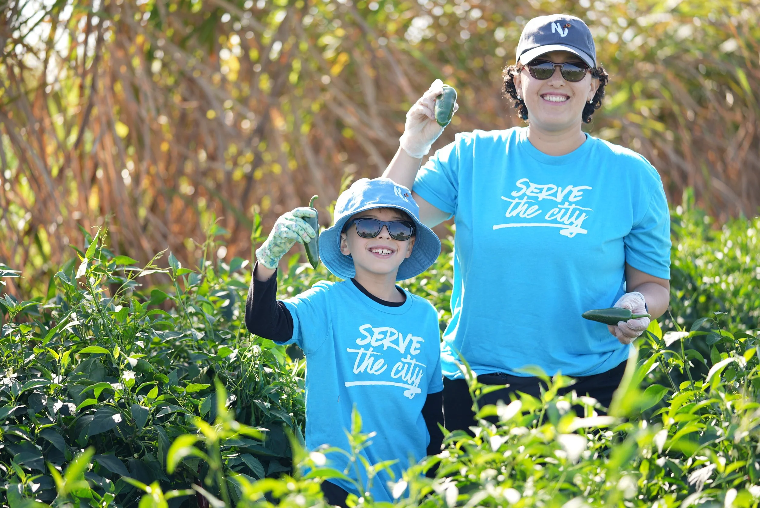 Woman and boy in a field of green plants holding small green peppers, both wearing blue T-shirts with the text 'SERVE the city', sunglasses, hats, and gloves, smiling and posing for the photo.