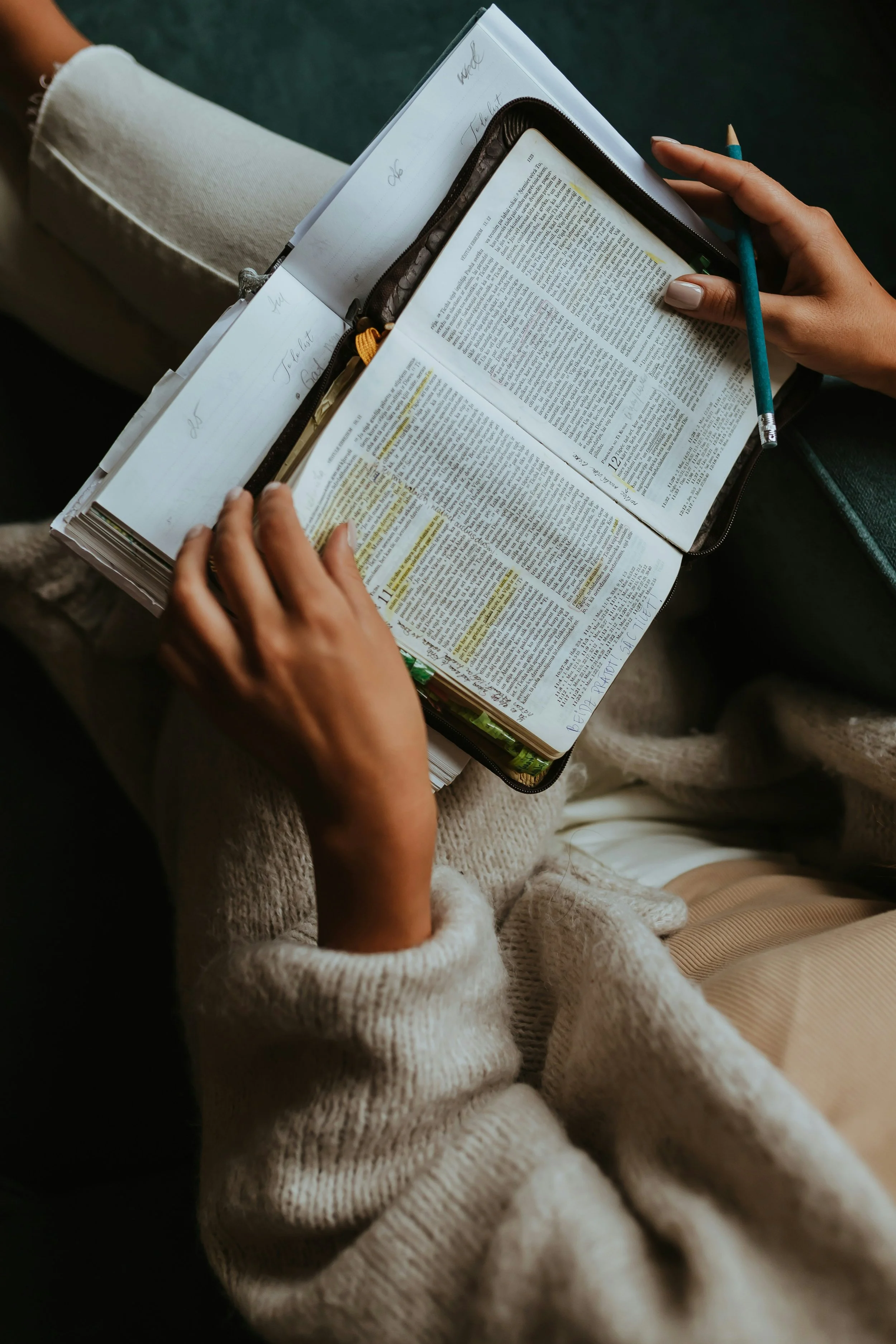 Close-up of a person holding an open book with highlighted text, sitting on a beige armchair with another person nearby wearing a cream-colored sweater.