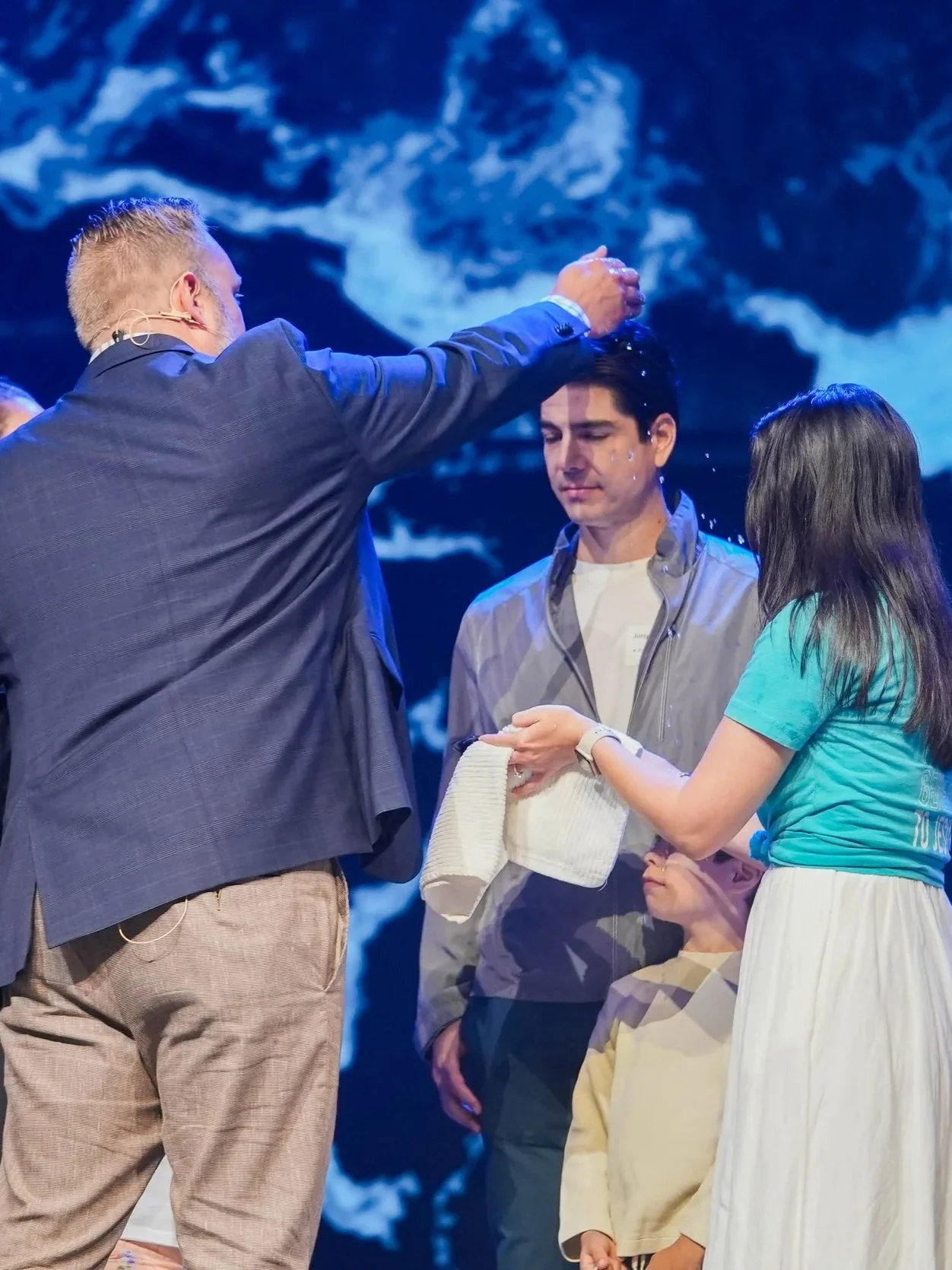 A man being baptized by a pastor in a church, with another girl holding a towel nearby, during a religious ceremony.