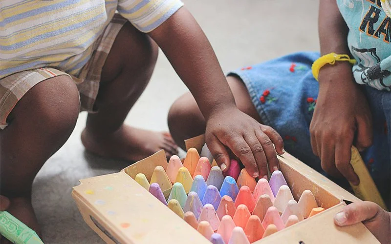Two children kneel on the floor and reach into a box of colorful chalks.