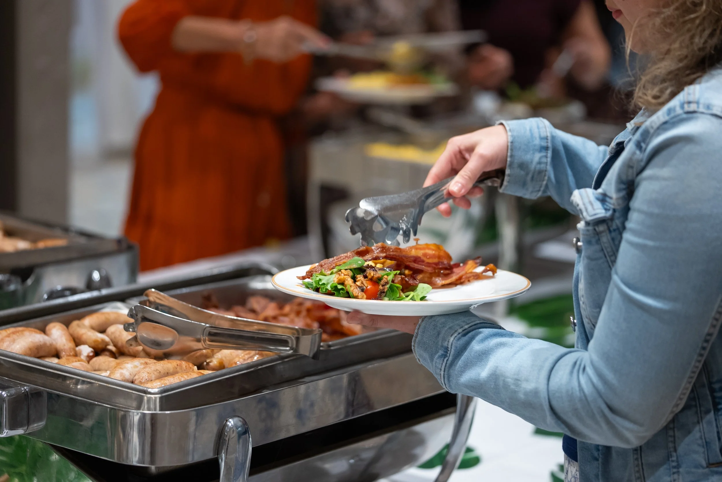 Person filling a plate with food from a buffet. The plate contains vegetables and meat. Other buffet trays with sausages and baked goods are visible in front.