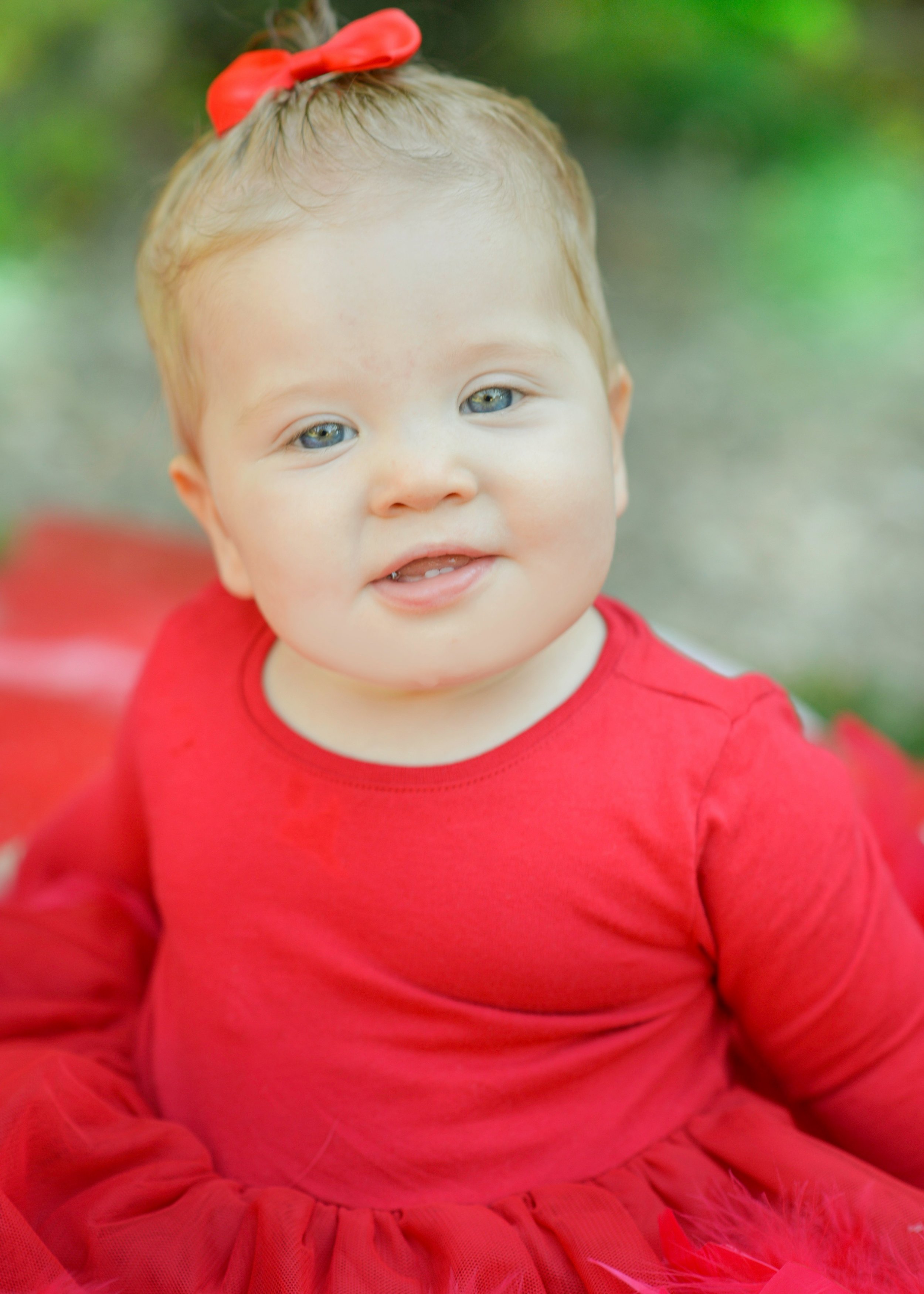 A young child with light skin, blonde hair, and blue eyes wearing a red bow in her hair and a red shirt, smiling outdoors with a blurred green background.