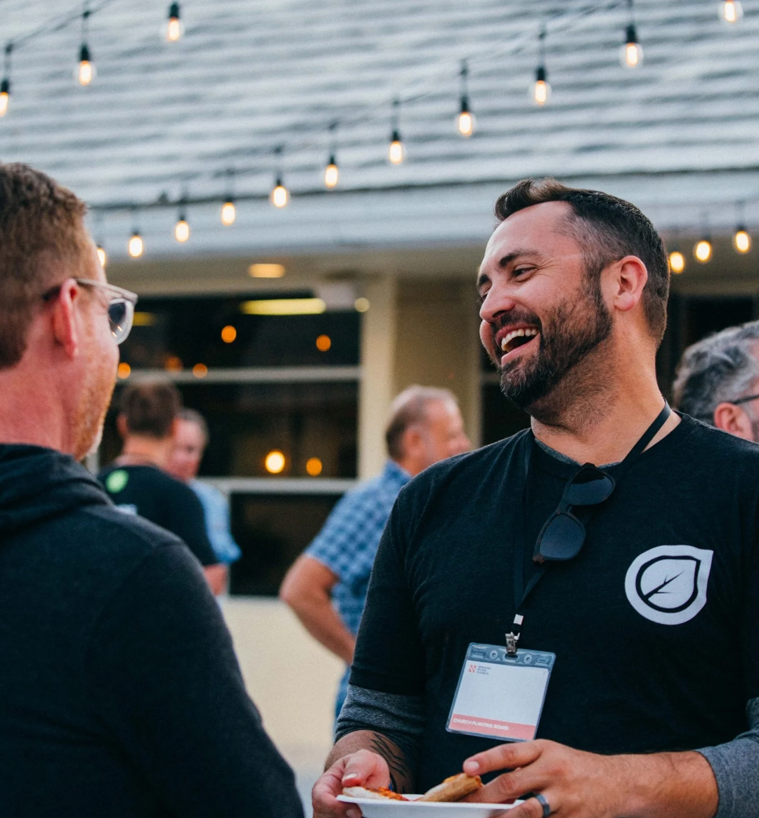 Two men smiling and talking at an outdoor event, one holding a plate of food, with string lights hanging overhead.