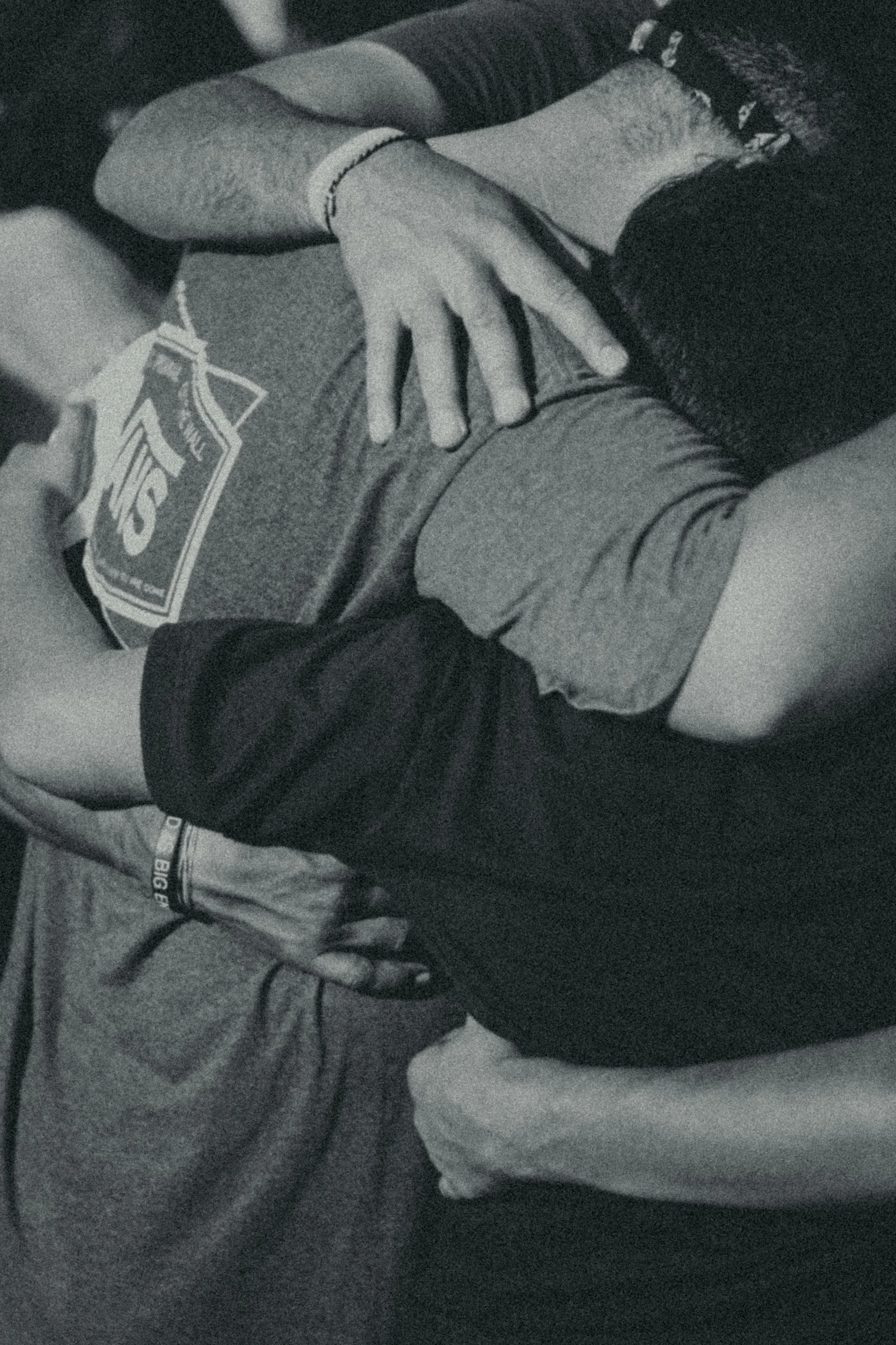 A black and white photo showing a close-up of two people lying on a surface, with one person's hand resting on the other's stomach. The person with the hand is wearing a wristband and a bracelet. The individuals are dressed casually, and the image conveys a sense of intimacy or closeness.