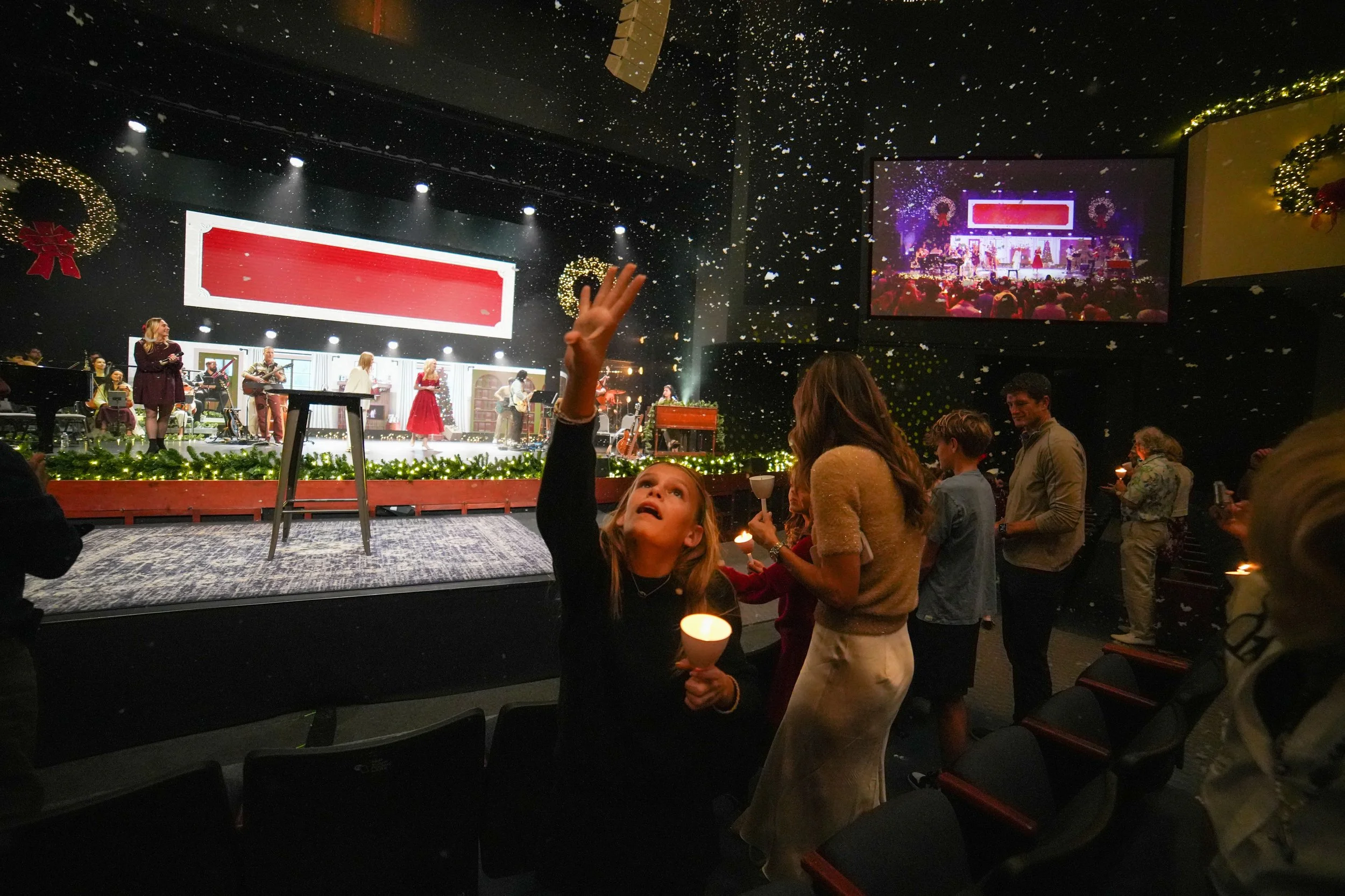 People attending a holiday concert or event, with a girl in the foreground reaching up while holding a candle, others holding candles, Christmas wreaths, and festive decorations, as a band performs on stage decorated with Christmas ornaments and lights.