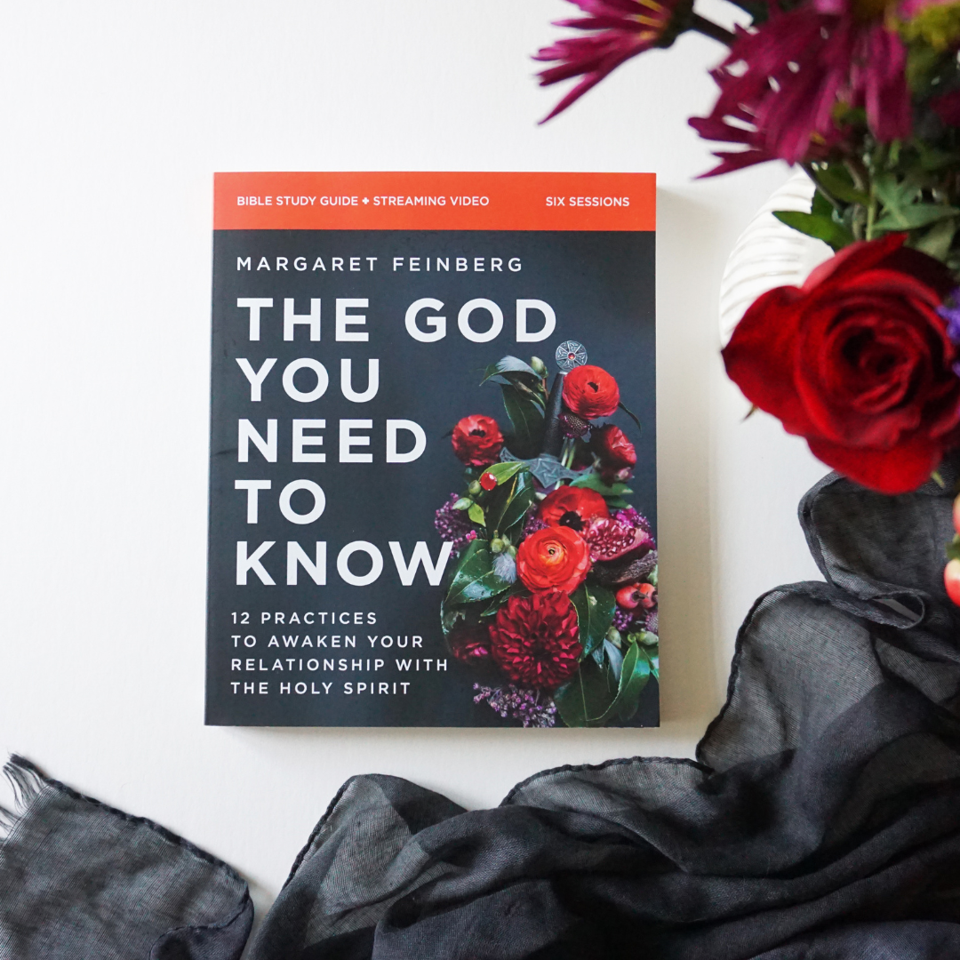 A book titled 'The God You Need to Know' by Margaret Feinberg, surrounded by a bouquet of red roses and dark fabric, on a white surface.