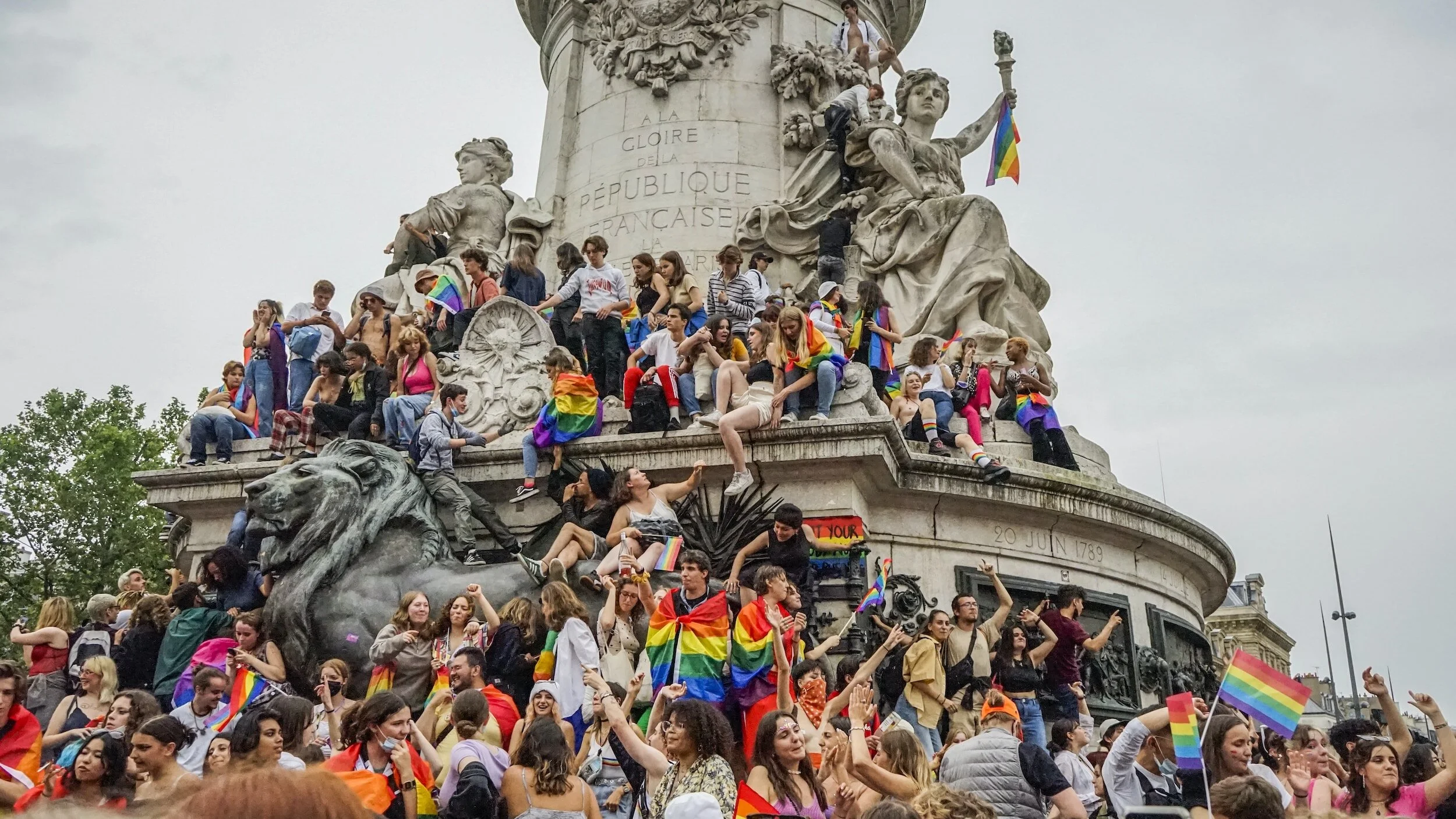 Crowd of people celebrating at a public monument with rainbow flags and rainbow clothing, during a Pride event.