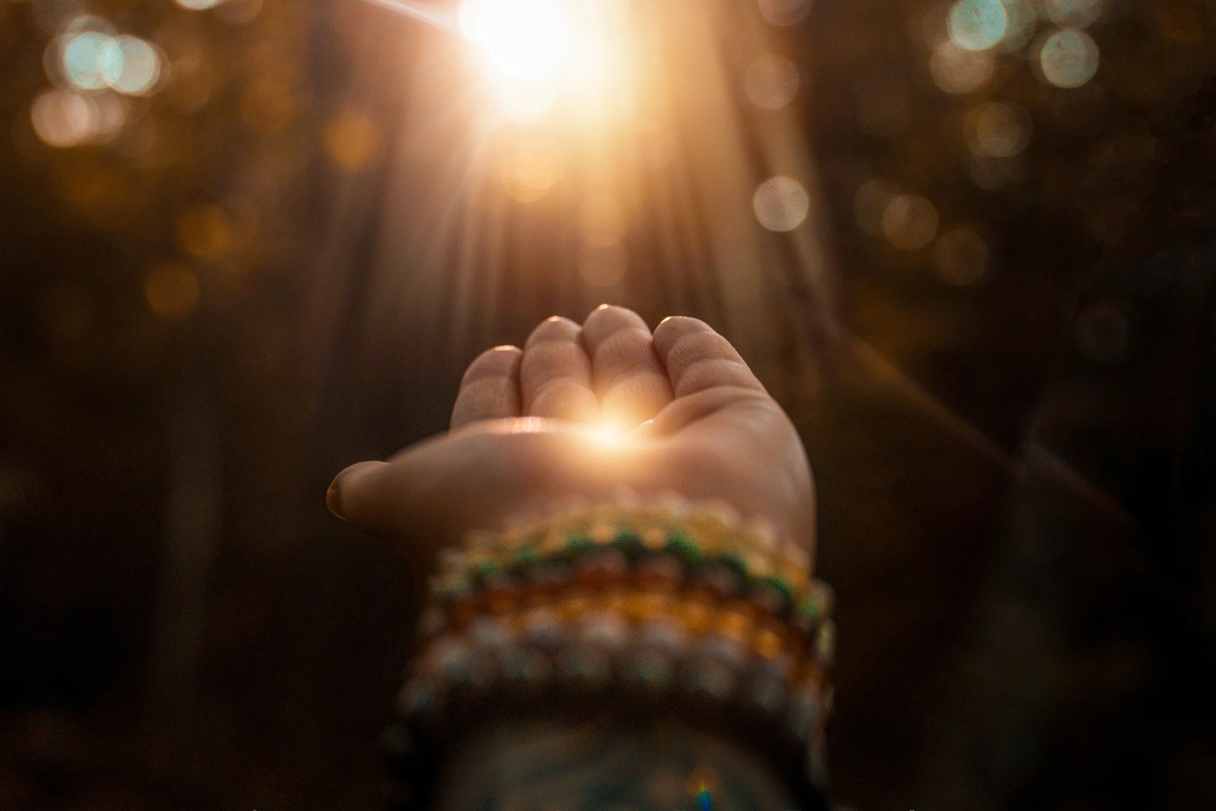 A person's hand reaching toward sunlight with trees in the background, surrounded by bokeh lights.