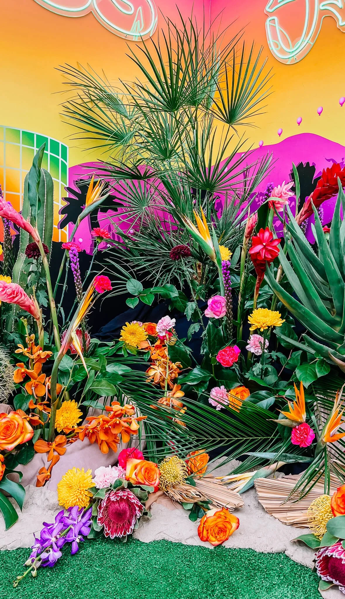 A portion of the large photo backdrop floral installation that shows the various bright colored flowers, succulents, a palm tree, palm fronds, and kinetic sand that were used in the design.